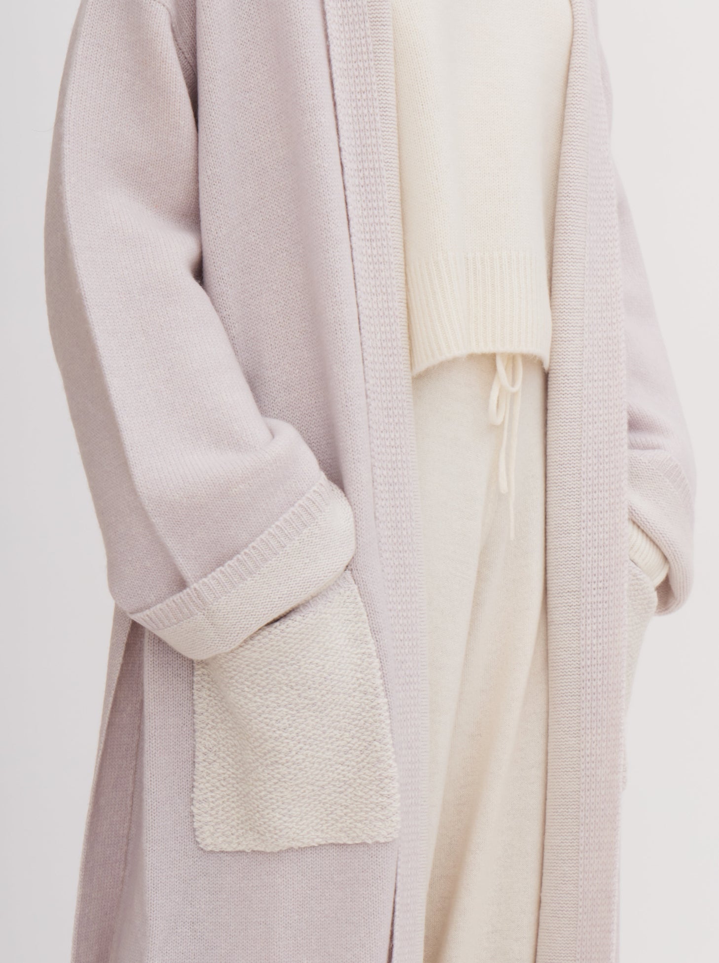 Close up detail showing lavender knitted cashmere long robe coat with wide sleeves and patch pockets, worn over white roll neck jumper and white cashmere pants.