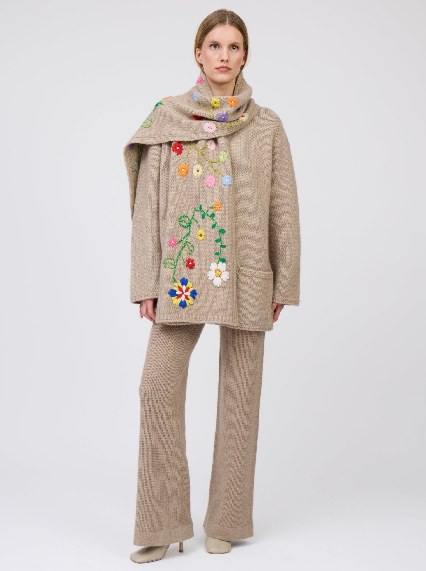 Front view model wearing light brown cashmere hip-length jacket with brightly coloured hand-embroidered scarf; worn with matching light brown knit pants.