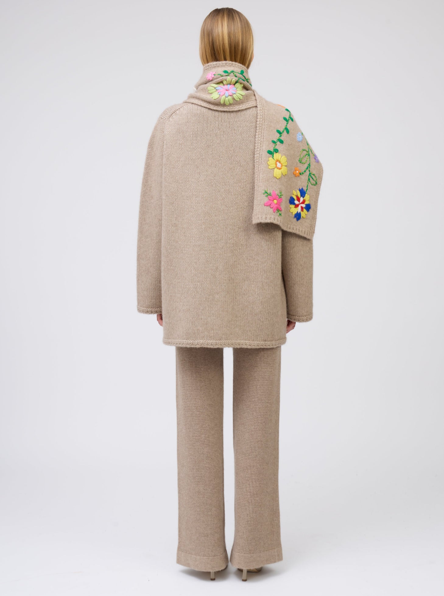 Back view model wearing light brown cashmere hip-length jacket with brightly coloured hand-embroidered scarf over one shoulder; worn with matching light brown knit pants.
