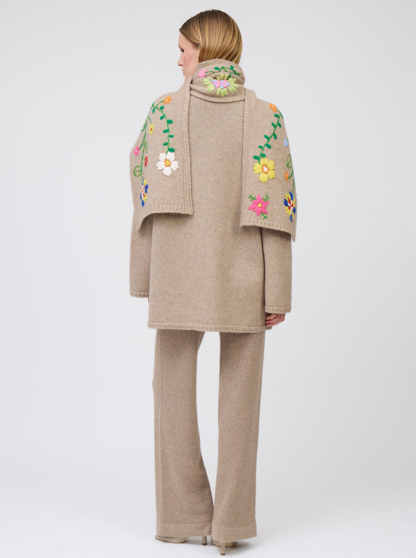Back view model wearing light brown cashmere hip-length jacket with brightly coloured hand-embroidered floral scarf thrown over both shoulders; worn with matching light brown knit pants.