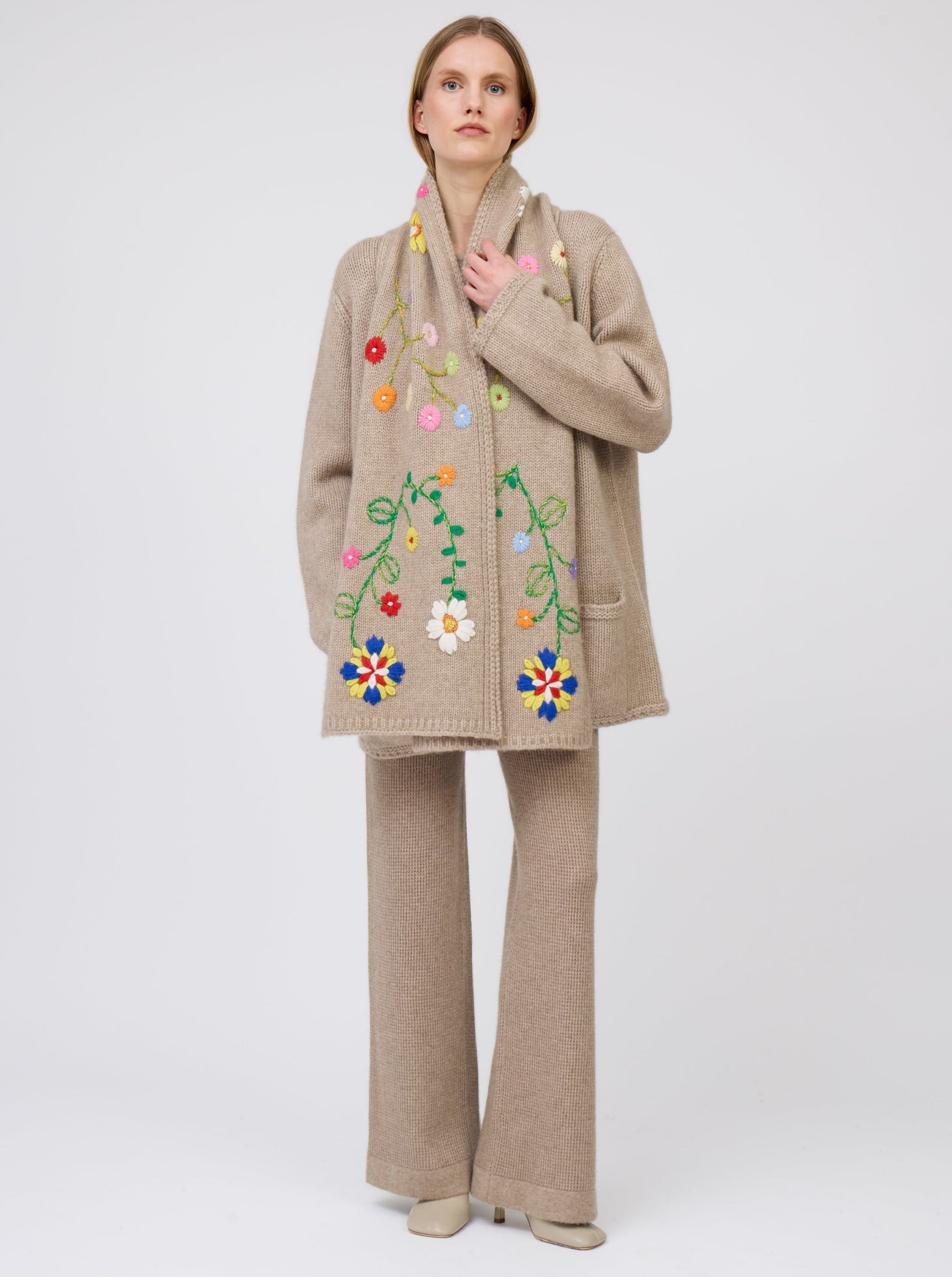 Front view model wearing light brown cashmere hip-length jacket with brightly coloured hand-embroidered scarf worn down the centre front of jacket; worn with matching light brown knit pants.