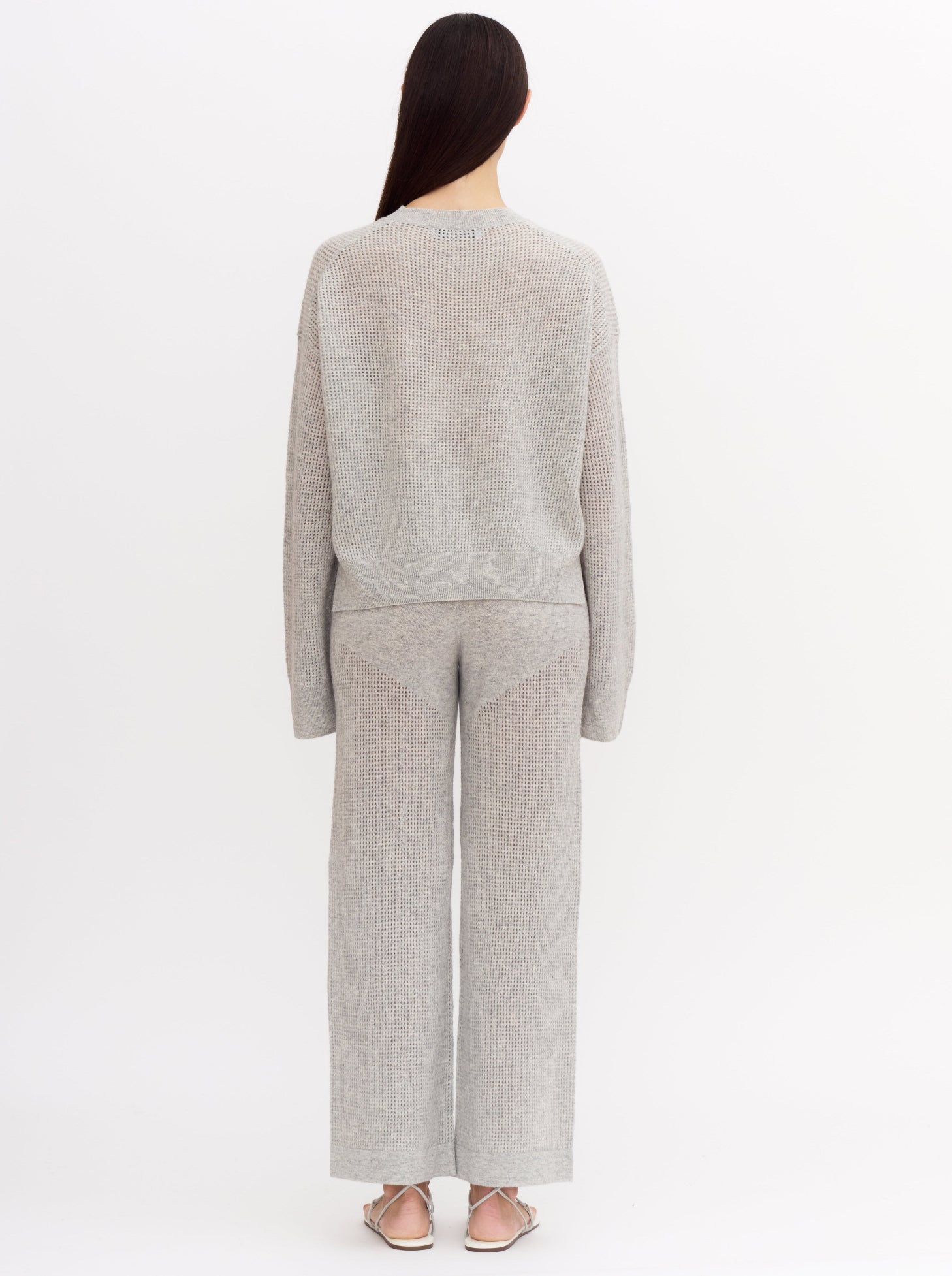Back view of model wearing light grey cashmere net knit cashmere long sleeve sweater with wide leg matching light grey cashmere pants.