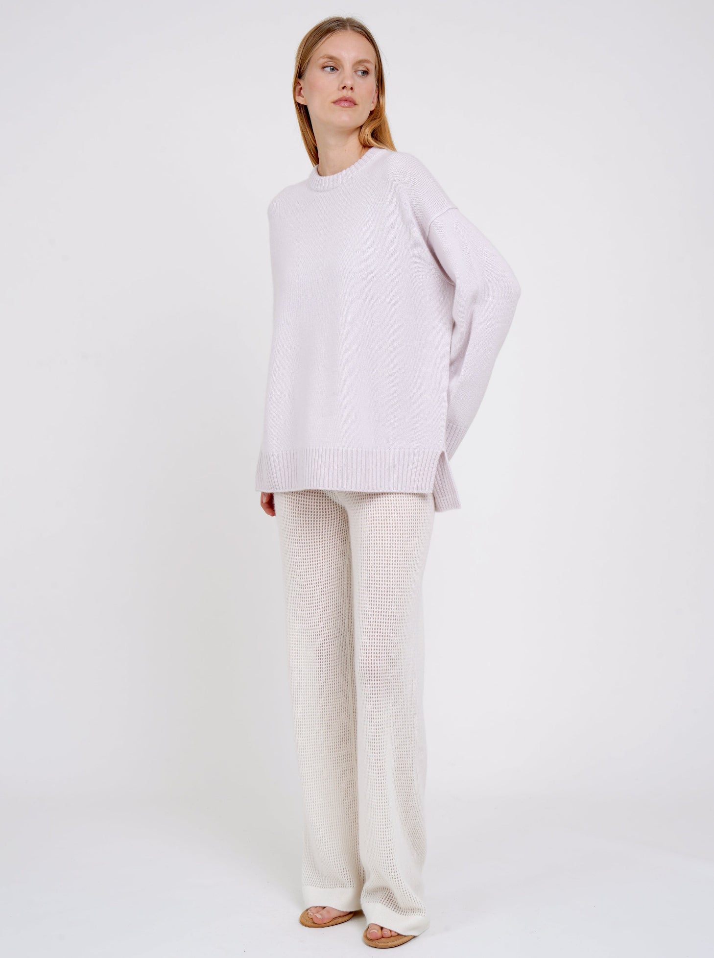 Standing model posing in pale purple long jumper with round neck, long sleeves and white cashmere knit pants.