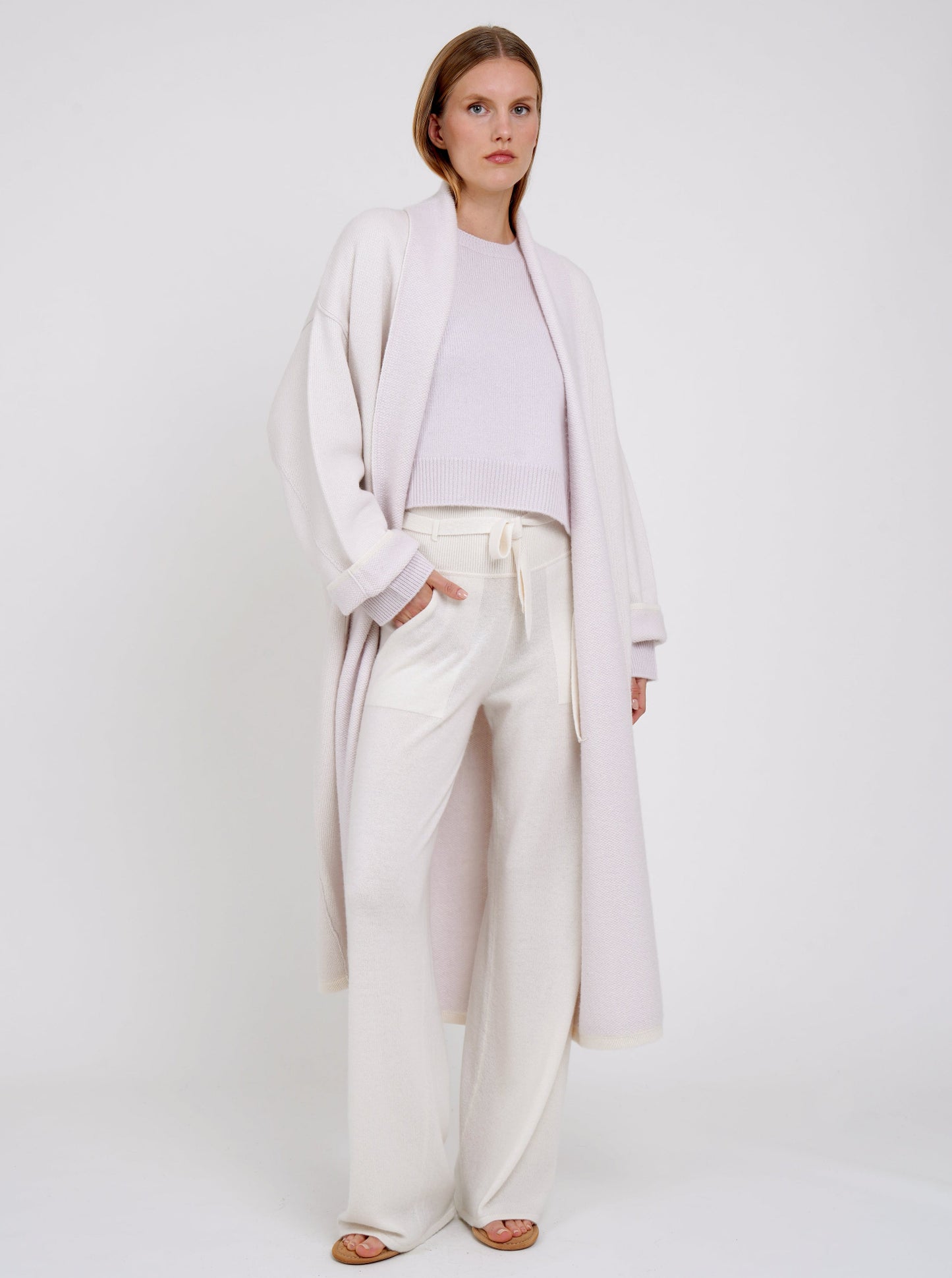 Model in lilac cashmere knit long coat with collar revere and turn back cuffs; worn with lilac cropped jumper and wide leg white cashmere pants.