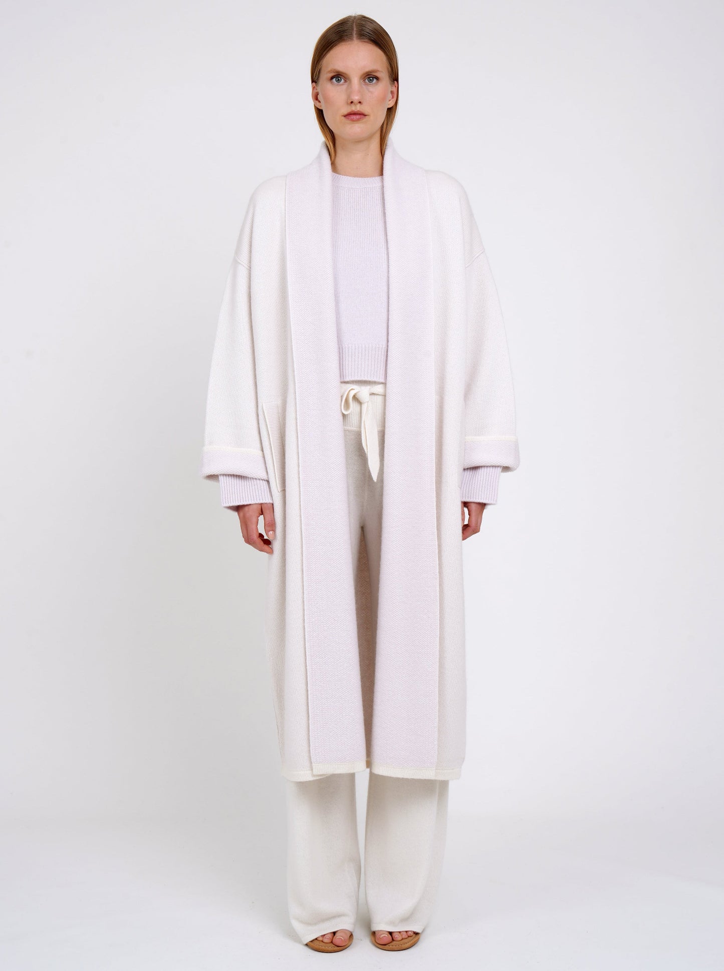 Front view model in lilac and white mix cashmere knit long coat with lilac collar revere and turn back cuffs; worn with lilac cropped jumper and wide leg, belted white cashmere pants.