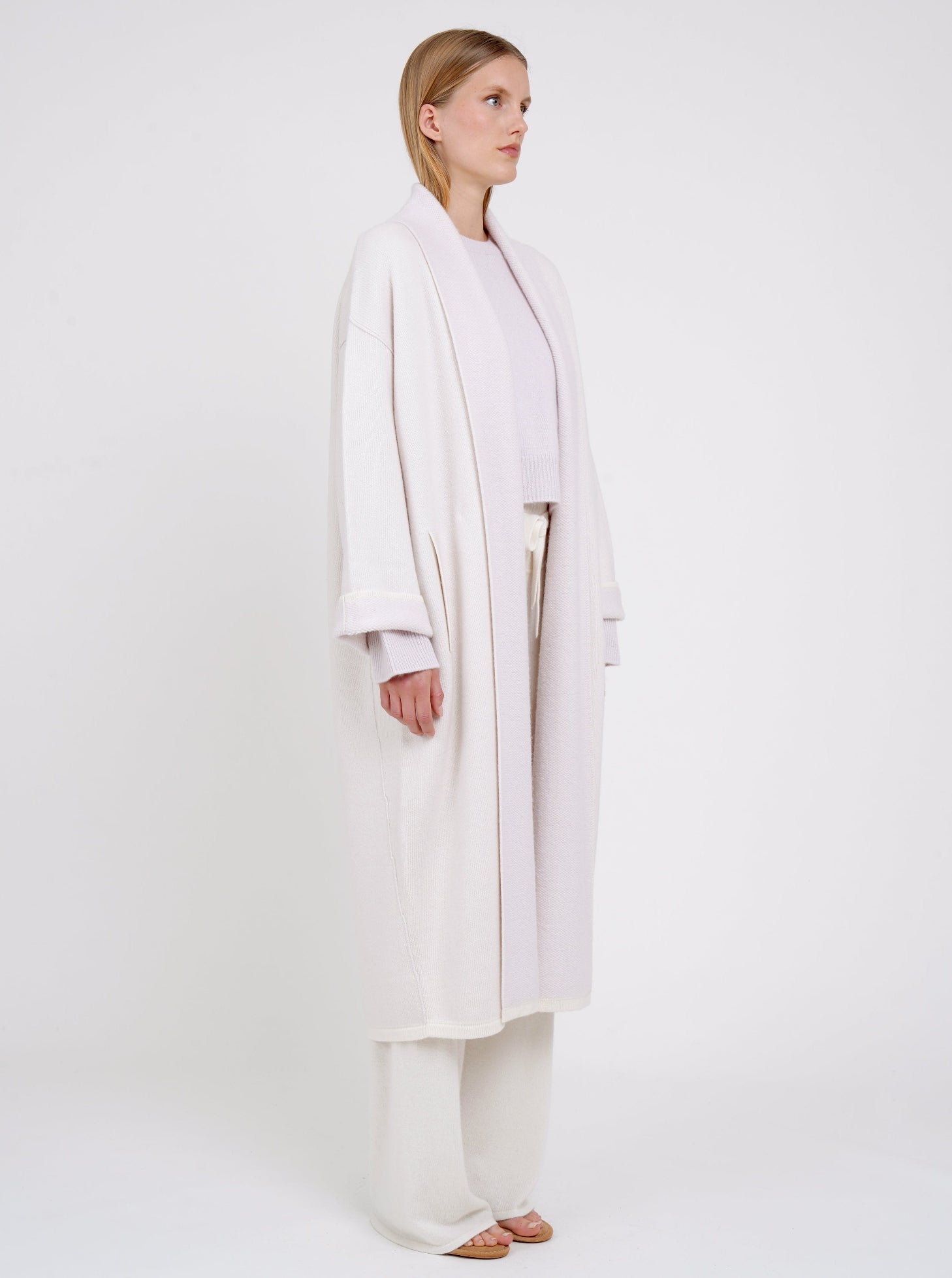 Side view of model in lilac and white mix cashmere knit coat with lilac collar revere and turn back cuffs; with lilac cropped jumper and wide leg white cashmere pants.