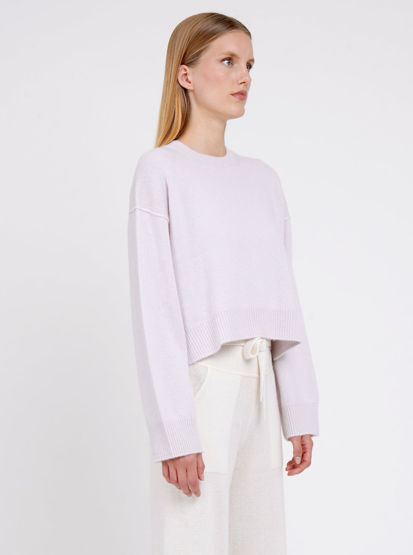 Side view close-up of model in lilac cashmere cropped jumper with round neck, long sleeves; worn with white cashmere, belted pants.