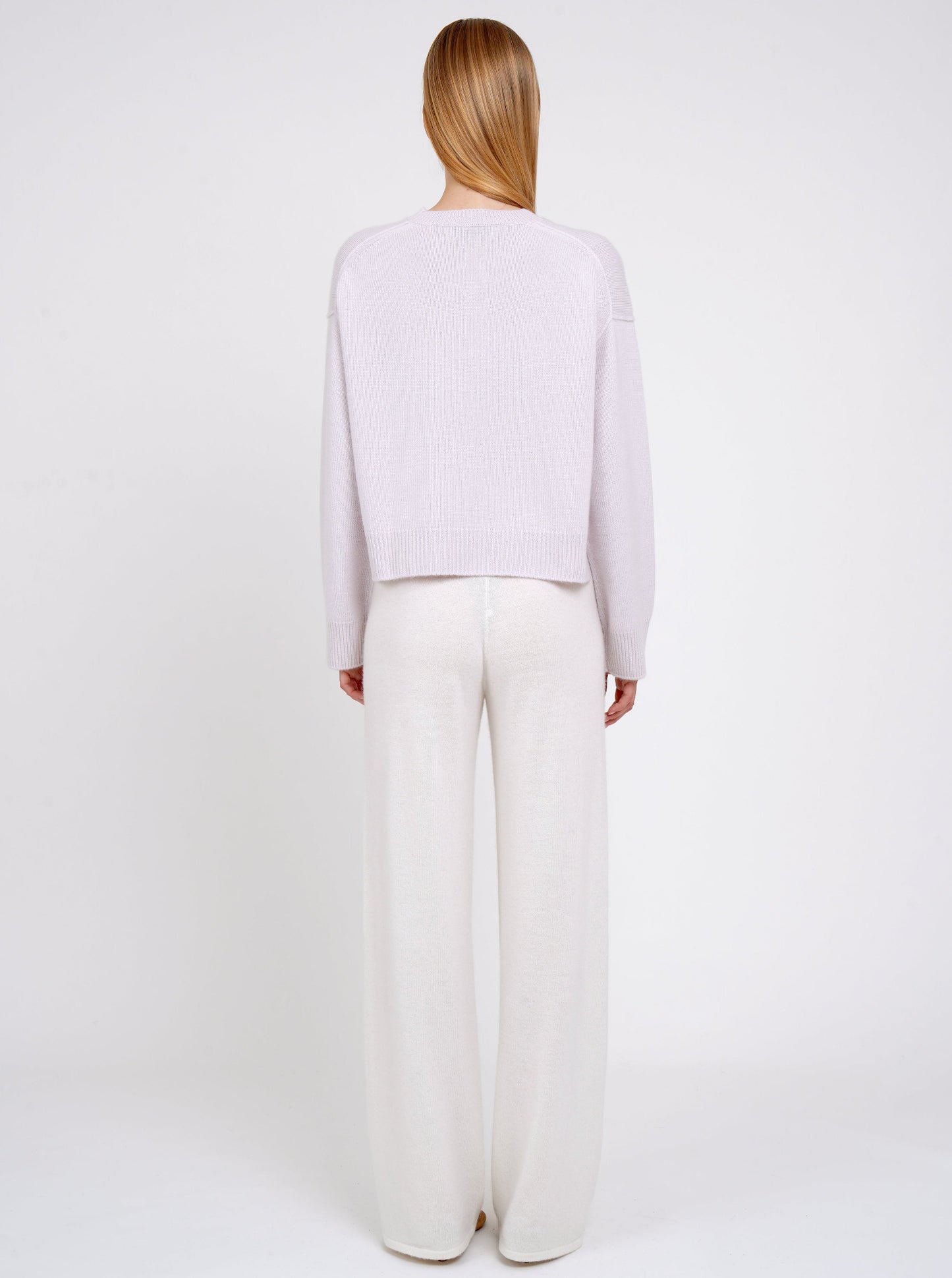 Back view of model in lilac cashmere cropped jumper with plain neck, long sleeves; worn with white cashmere, wide leg pants.