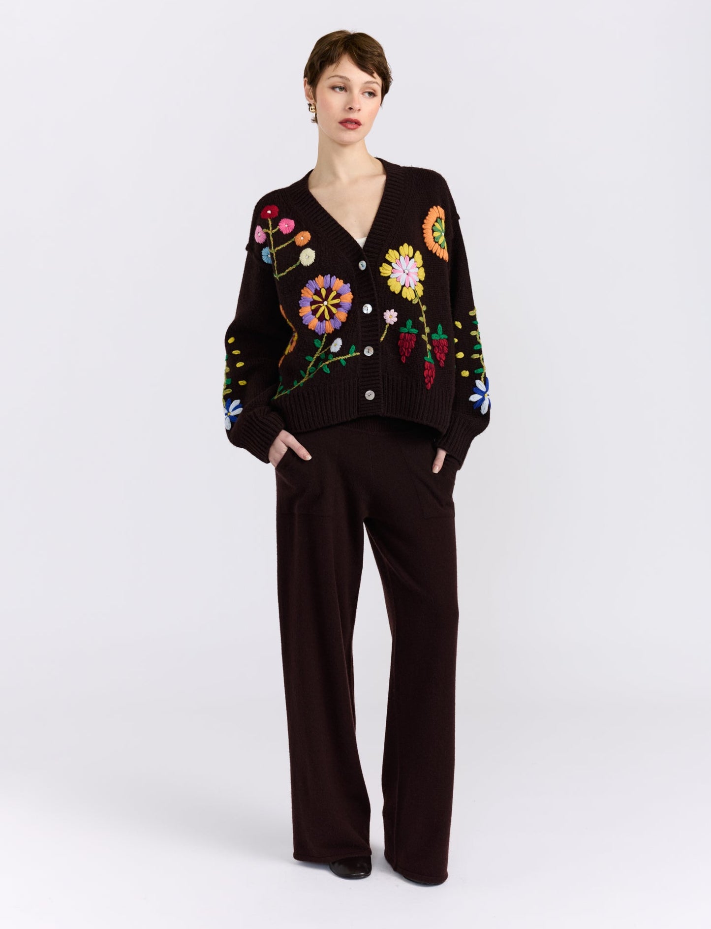 A model stands against a light gray studio background wearing a dark brown knit cardigan with colorful floral embroidery and matching dark brown wide-leg trousers.