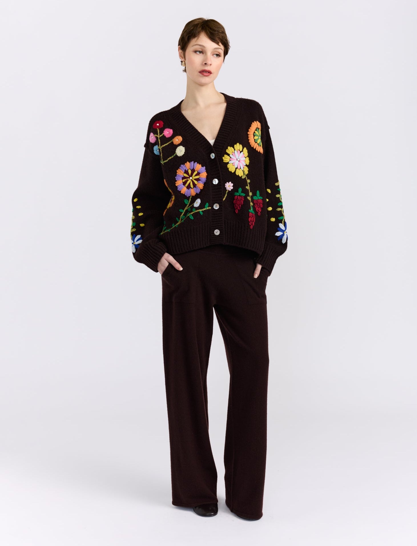 A model stands against a light gray studio background wearing a dark brown knit cardigan with colorful floral embroidery and matching dark brown wide-leg trousers.