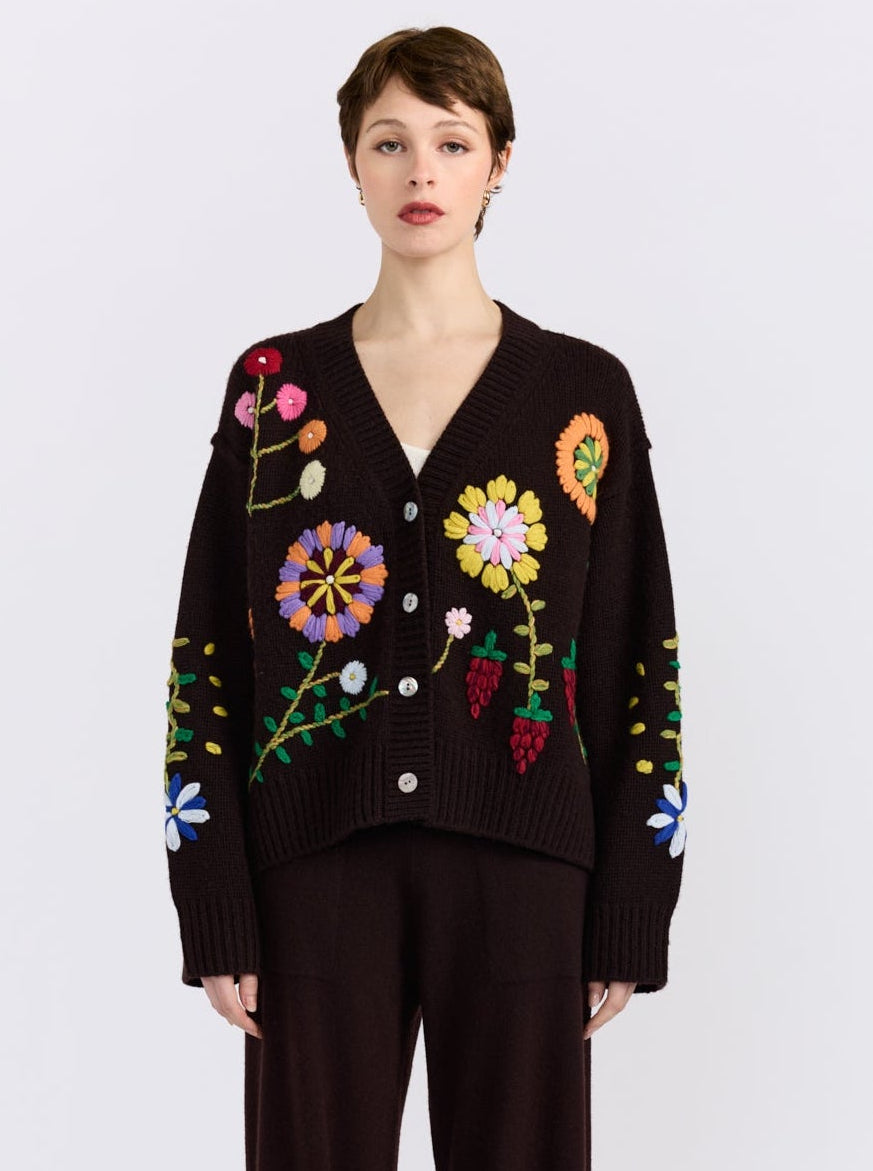 A model wearing a dark brown knit cardigan sweater with colorful embroidered flowers and leaves, paired with dark brown trousers, against a light gray background.