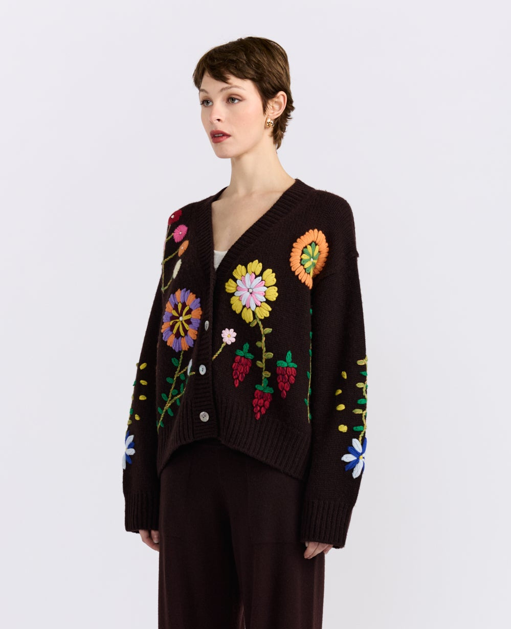 A model wearing a dark brown knit cardigan sweater with colorful embroidered flowers and leaves, paired with dark brown trousers, against a light gray background.