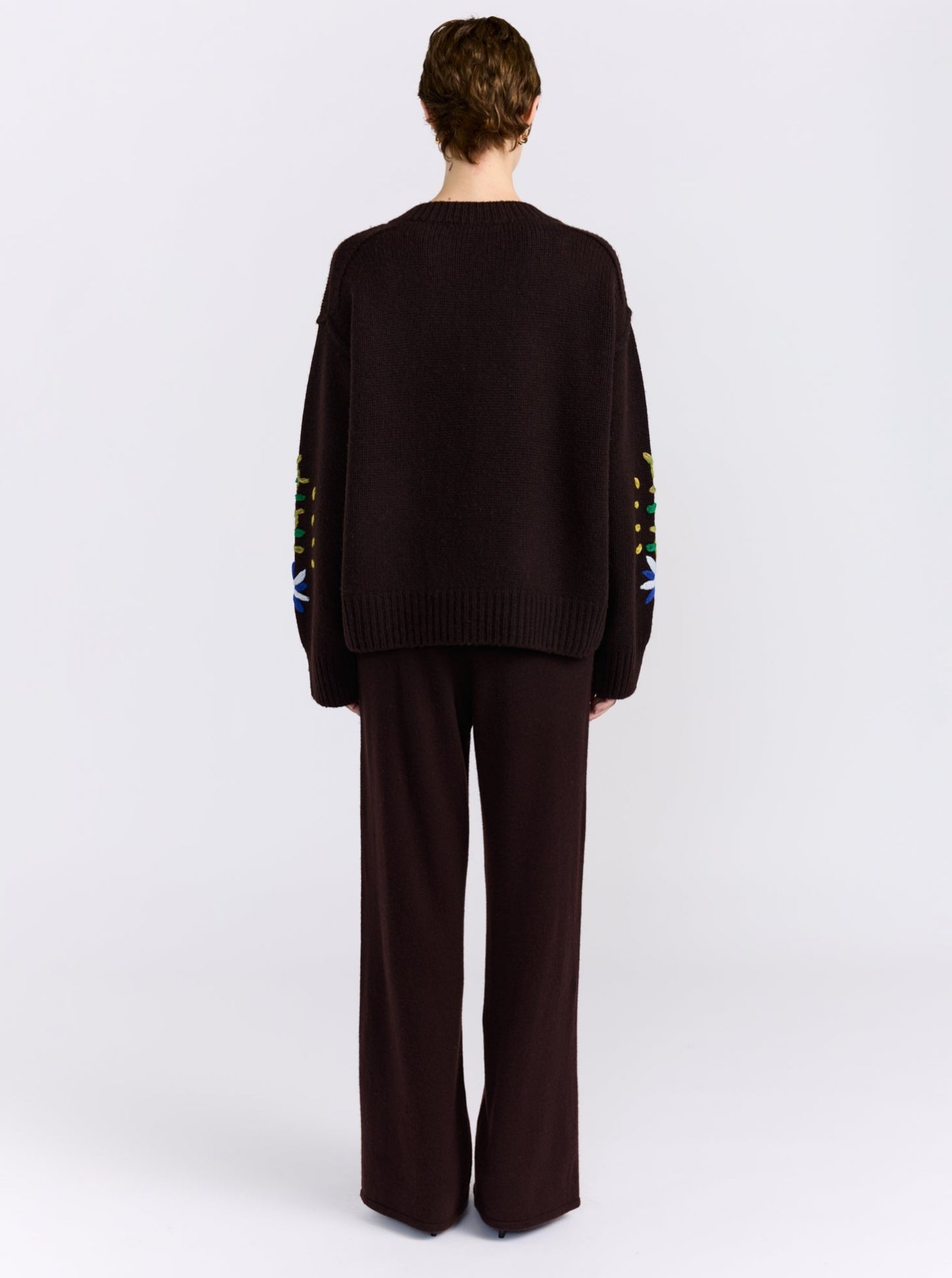 A model wearing a dark brown knit cardigan sweater with colorful embroidered flowers and leaves, paired with dark brown trousers, against a light gray background.