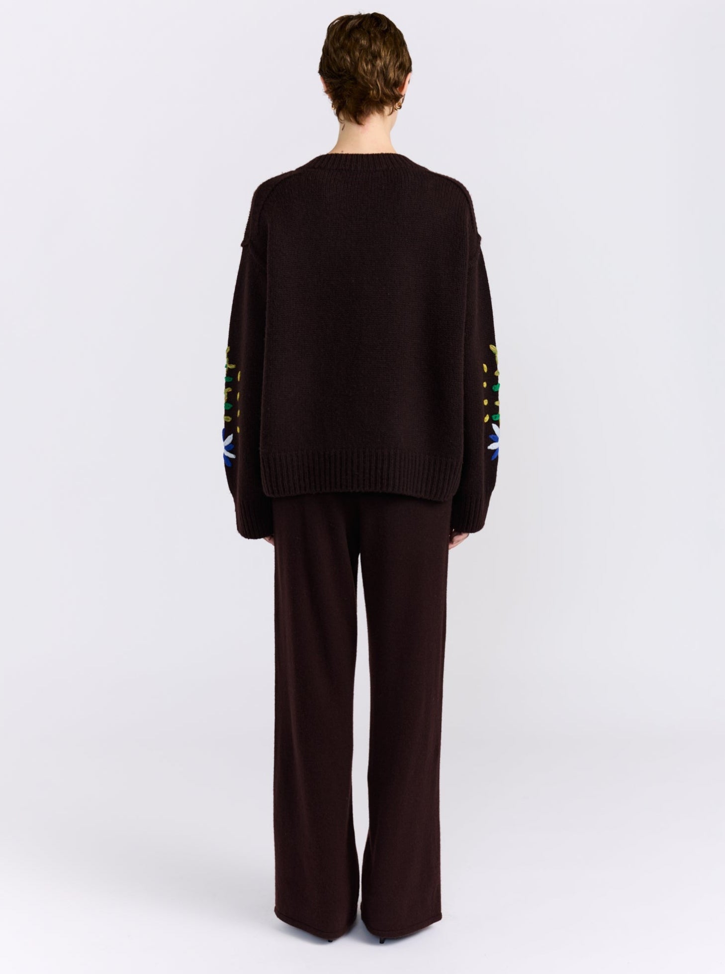 A model wearing a dark brown knit cardigan sweater with colorful embroidered flowers and leaves, paired with dark brown trousers, against a light gray background.