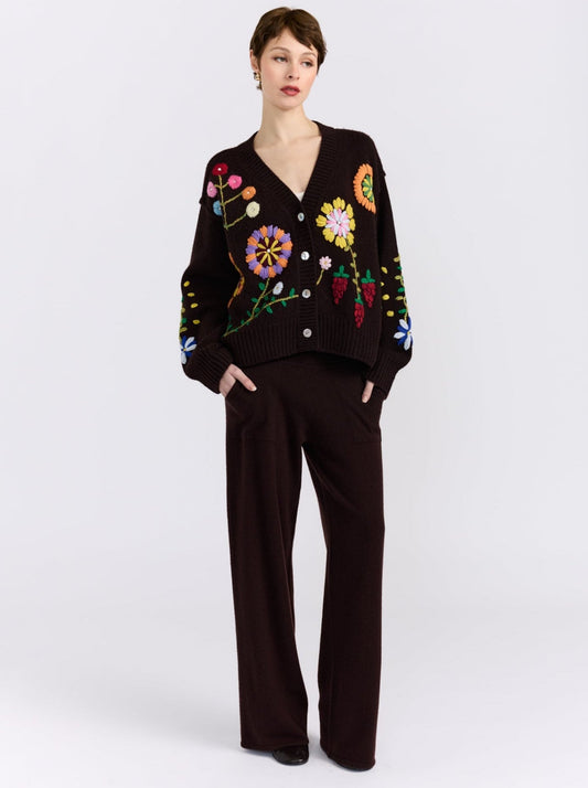A model stands against a white studio background wearing a dark brown knit cardigan with colorful floral embroidery and matching dark brown wide-leg trousers.