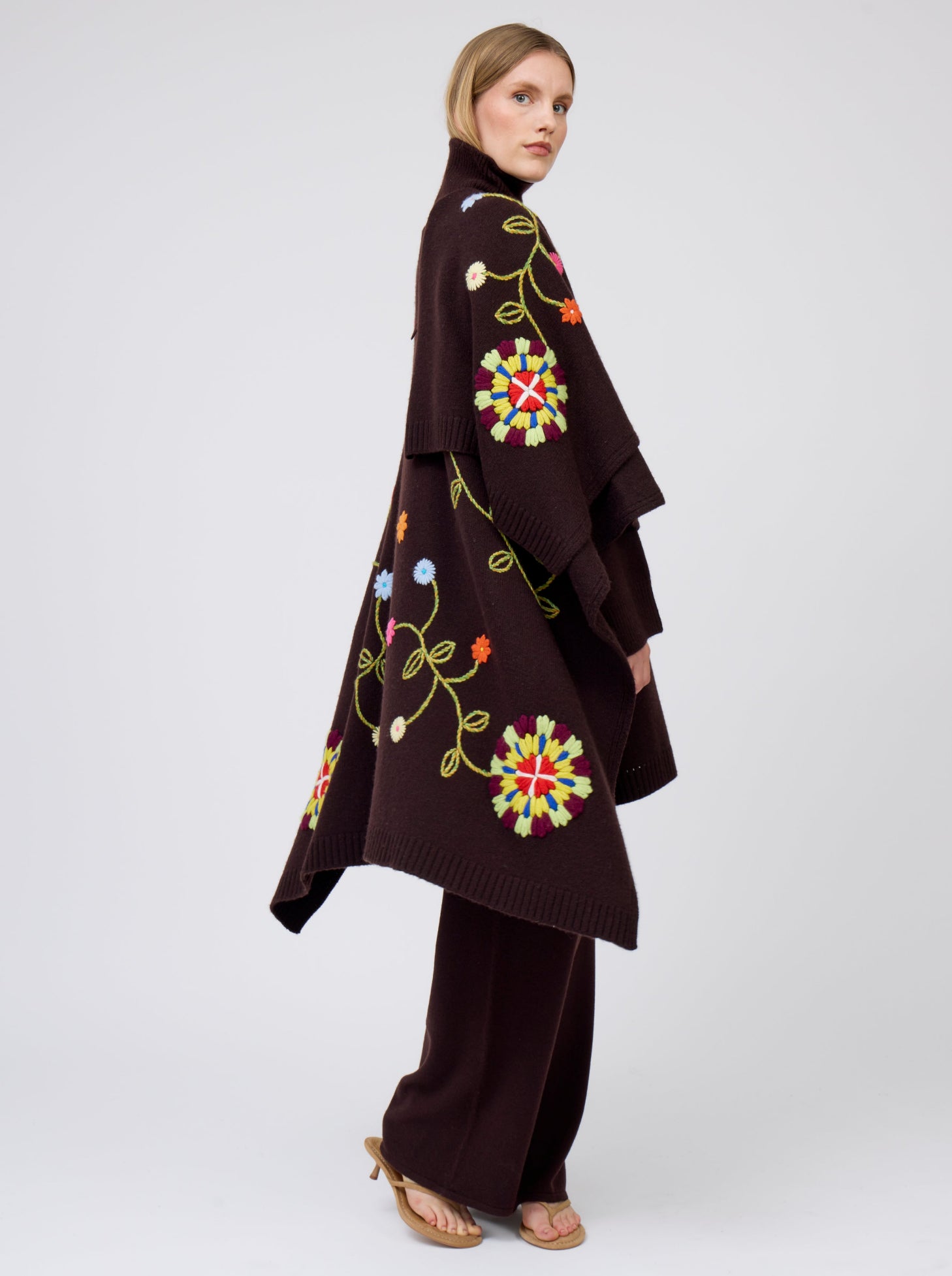 Moving shot of model showing flow of mahogany brown cashmere mid-length wrap with brightly coloured hand-embroidery; worn with matching brown jumper and brown pants.