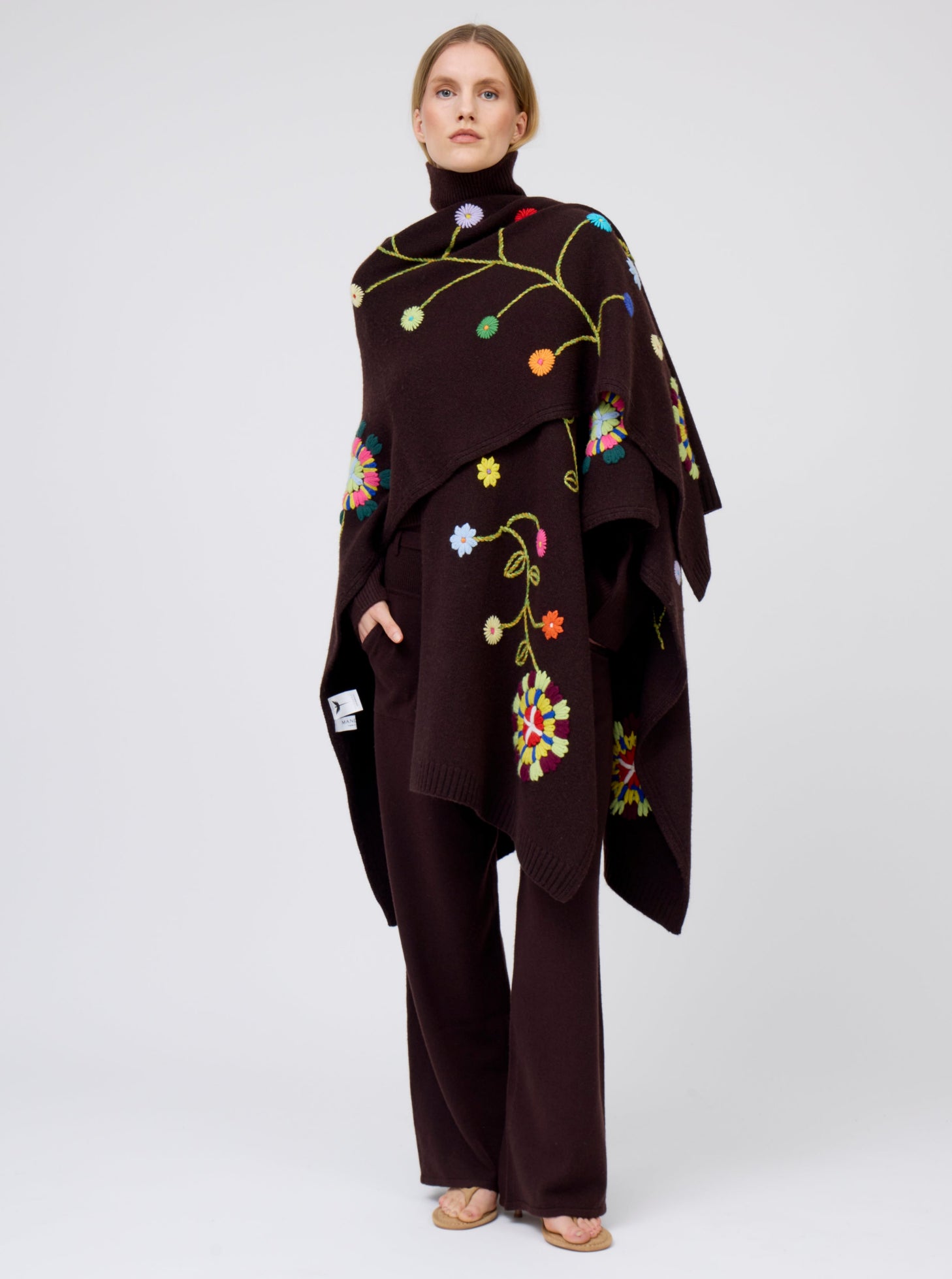 Model wears mahogany brown cashmere mid-length wrap with brightly coloured floral hand-embroidery, with one side thrown over shoulder; worn with matching brown jumper and brown pants.
