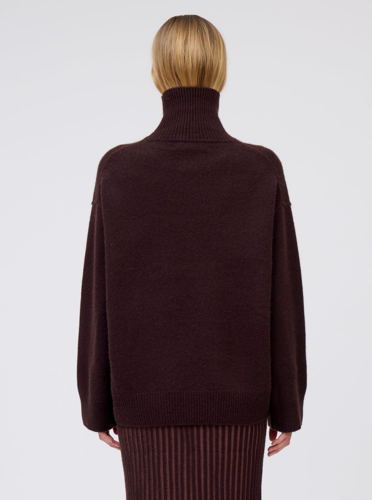 Close-up back view of model wearing mahogany brown cashmere oversized roll neck jumper with generous long sleeves and high-low hem.