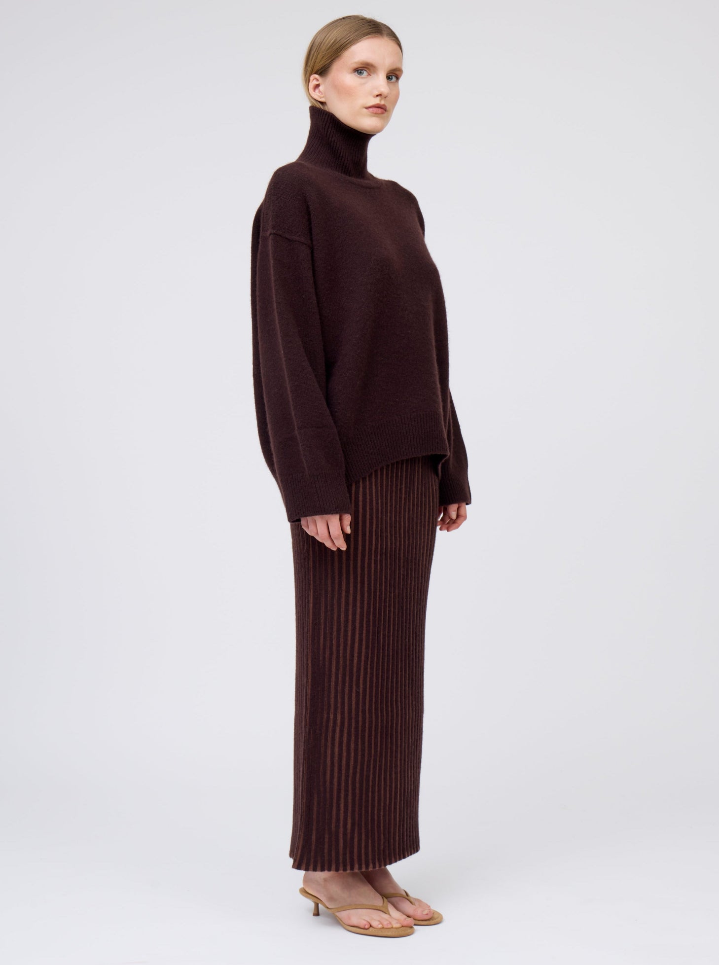 Model wears oversized mahogany brown cashmere roll neck jumper with generous long sleeves; worn with brown vertical stripe slim maxi skirt.