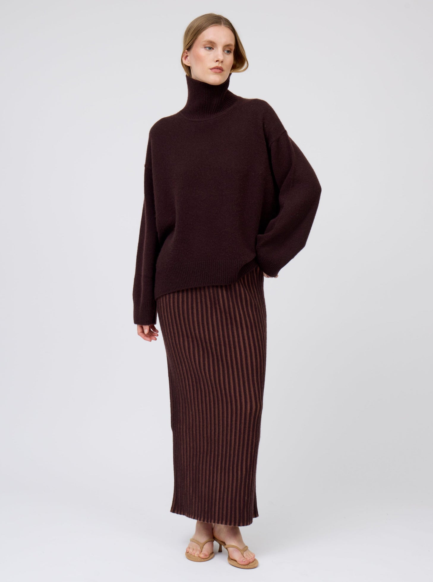 Model wears dark brown cashmere oversized roll neck sweater with generous long sleeves and high-low hem; worn with brown vertical stripe slim maxi skirt.