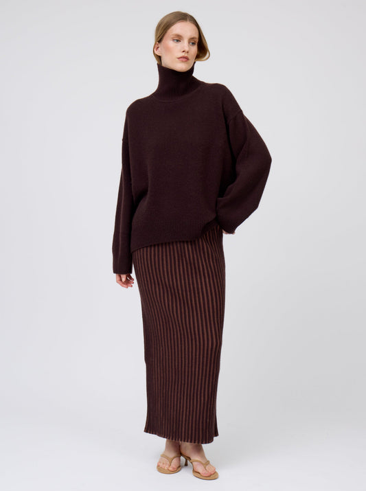 Model wears dark brown cashmere oversized roll neck sweater with generous long sleeves and high-low hem; worn with brown vertical stripe slim maxi skirt.