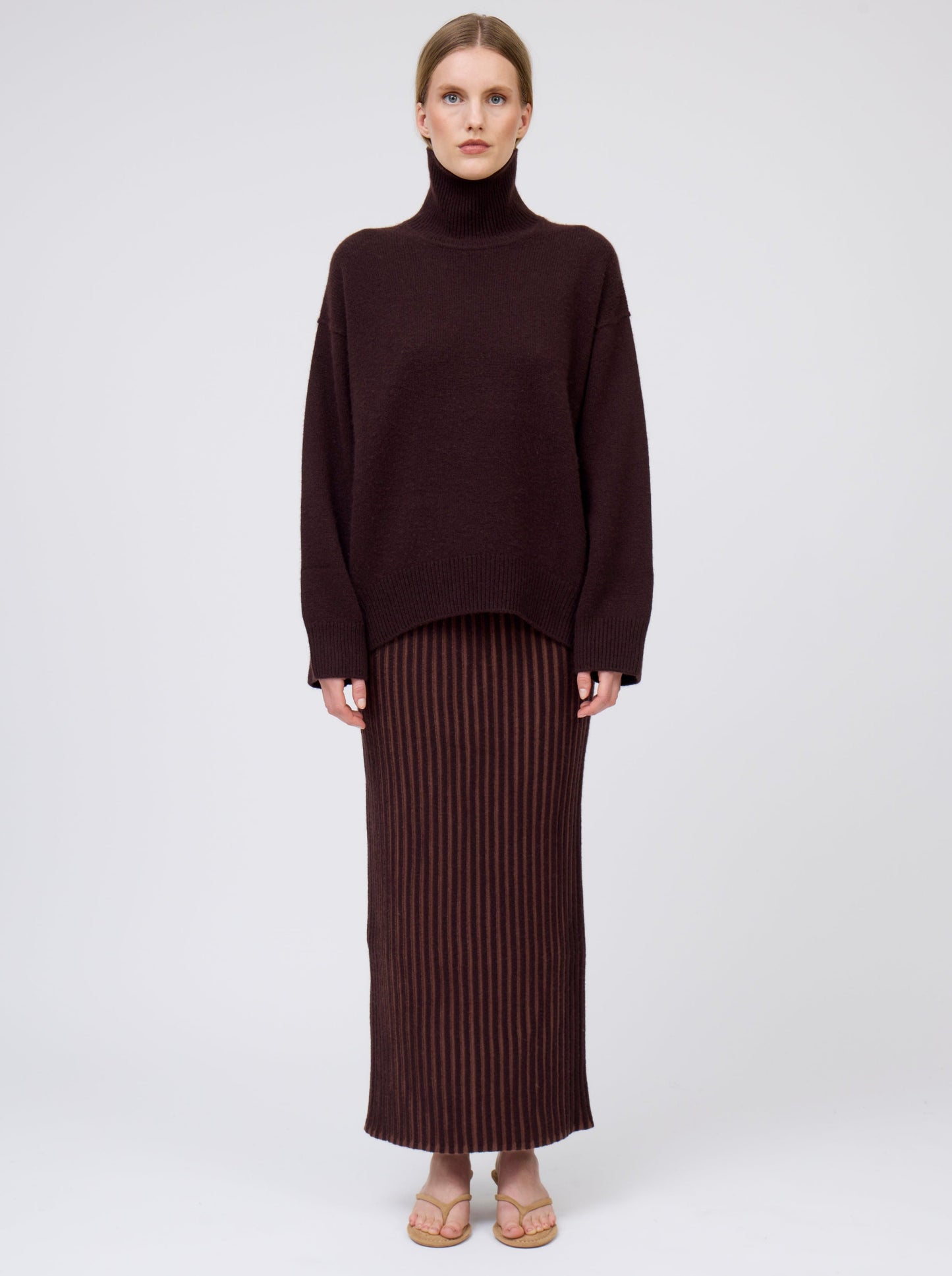 Front view of model wearing mahogany brown cashmere oversized roll neck sweater with generous long sleeves and high-low hem; worn with brown vertical stripe straight cut maxi skirt.