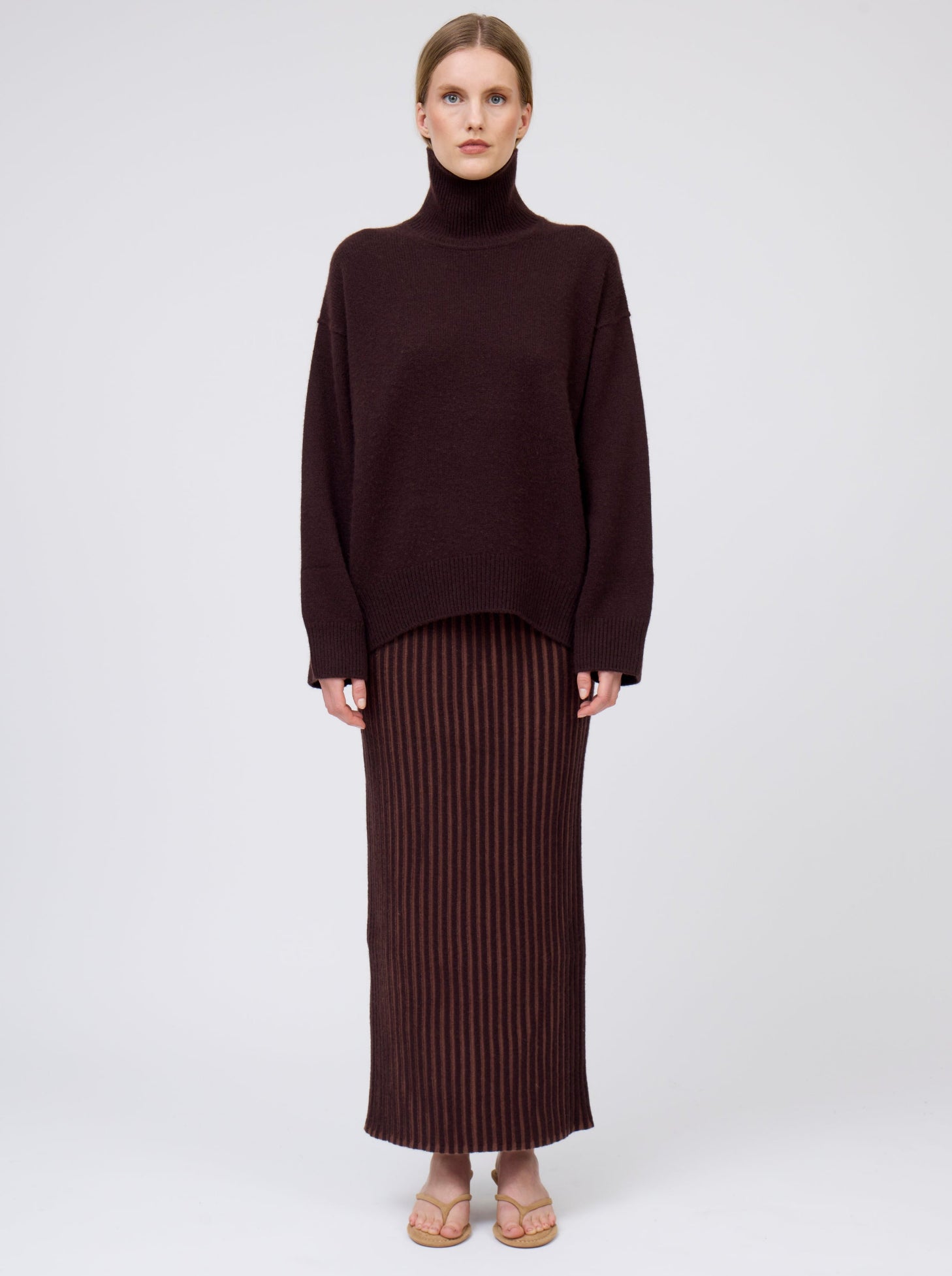 Front view of model wearing mahogany brown cashmere oversized roll neck sweater with generous long sleeves and high-low hem; worn with brown vertical stripe straight cut maxi skirt.