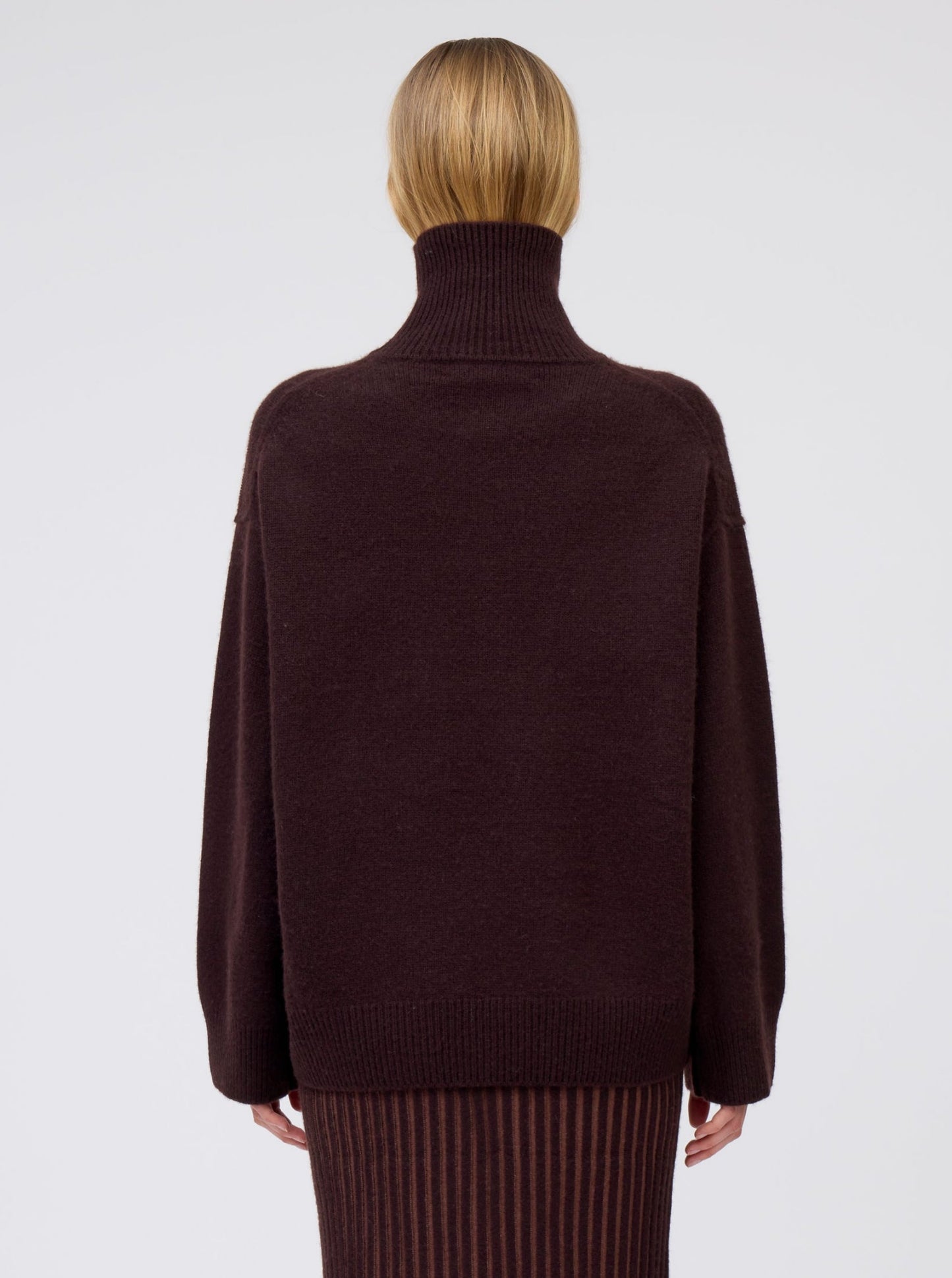 Close-up back view of model wearing mahogany brown cashmere oversized roll neck sweater with generous long sleeves and high-low hem.