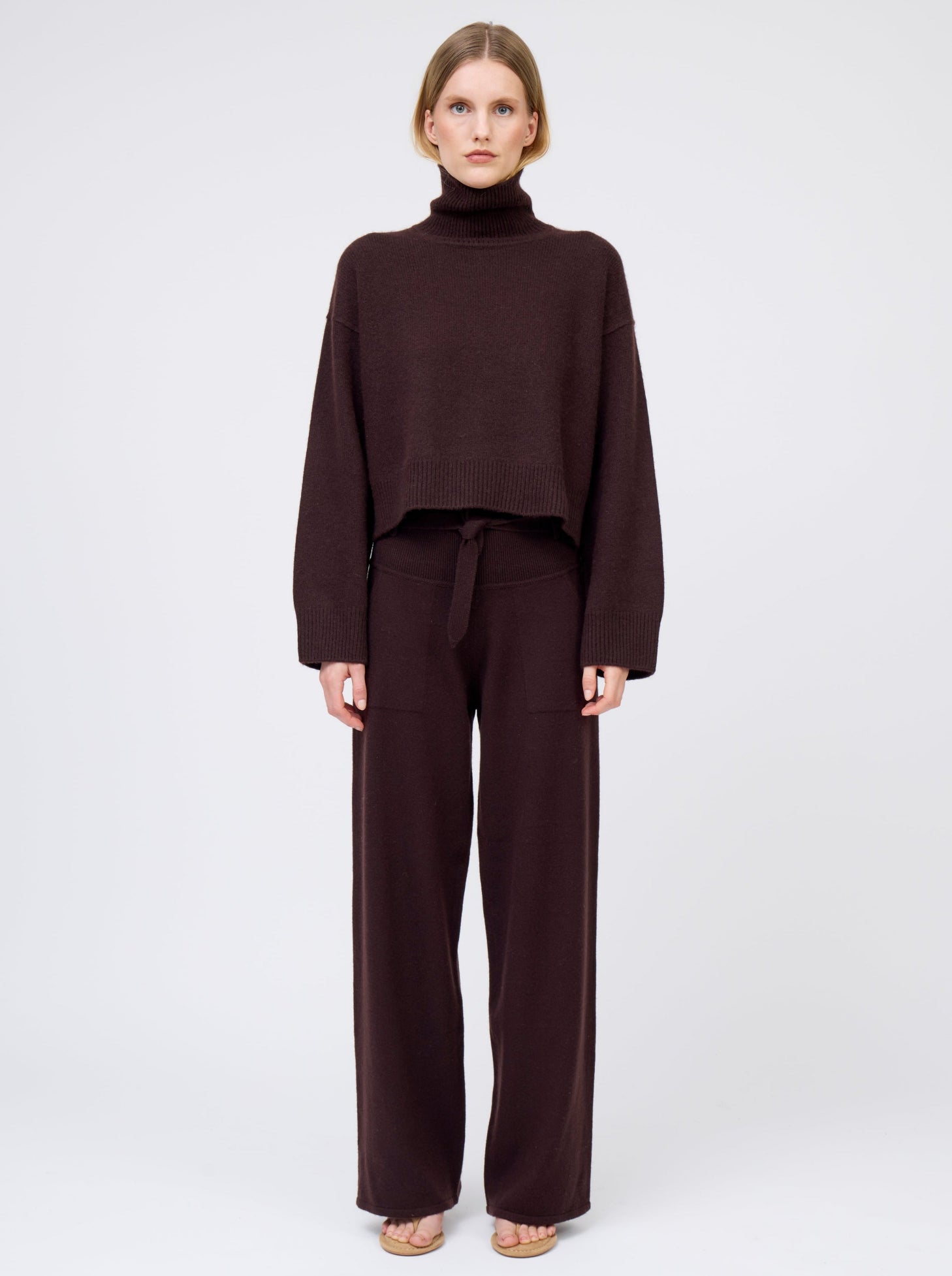 Front view of model wearing dark brown cashmere cropped length roll neck jumper with generous long sleeves and high-low hem; worn with  matching brown cashmere wide leg pants.