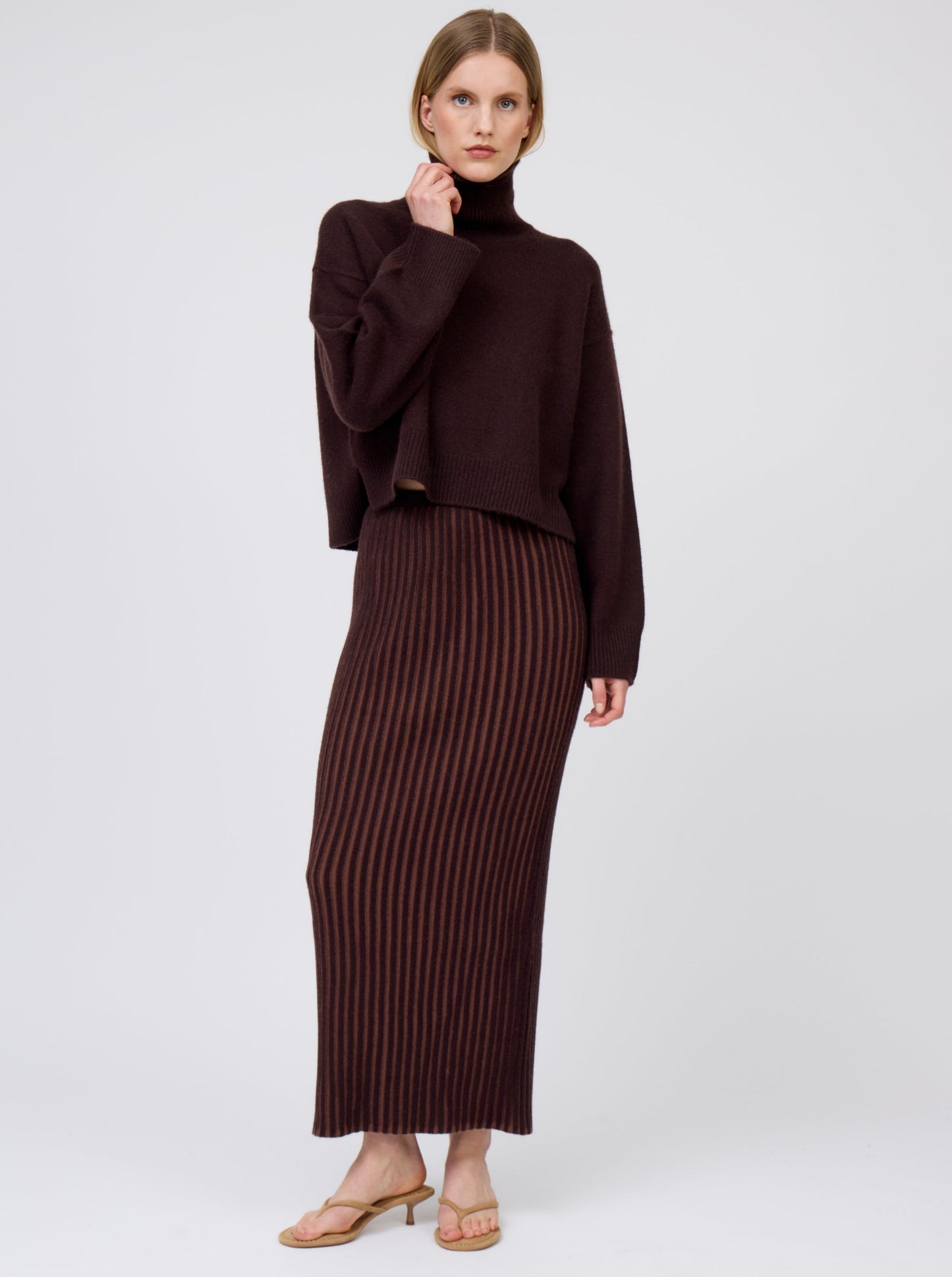 Model wears mahogany brown cashmere, cropped length, roll neck sweater with generous long sleeves and high-low hem; worn with brown vertical stripe slim tube skirt.