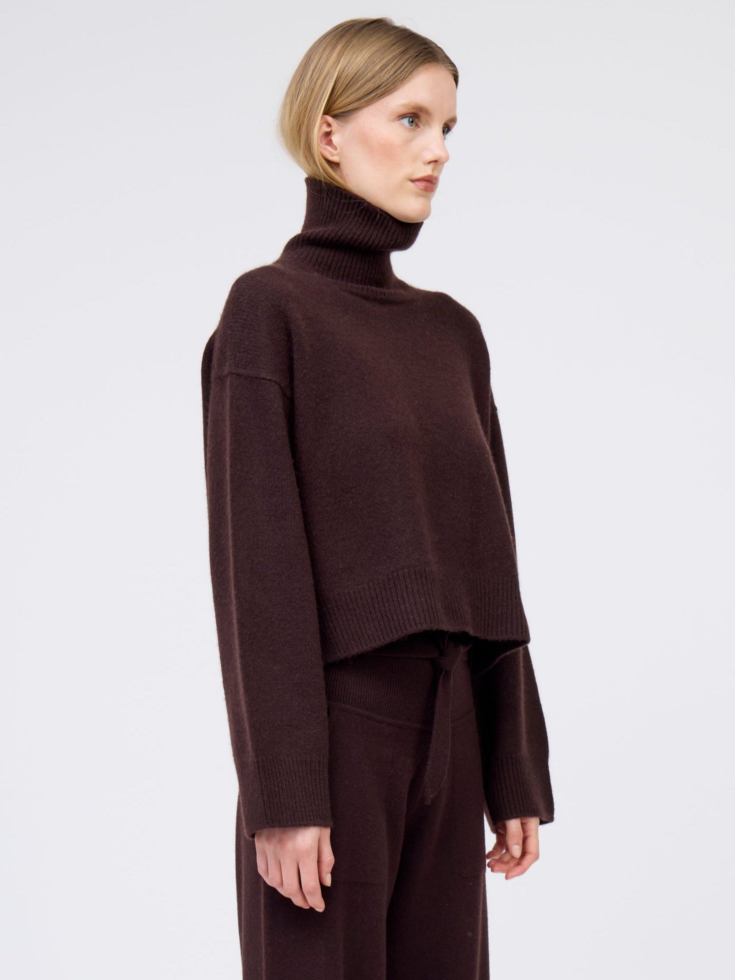 Close-up of model wearing mahogany brown cashmere cropped length, roll neck, sweater with generous long sleeves and high-low hem; worn with  matching brown cashmere belted pants.