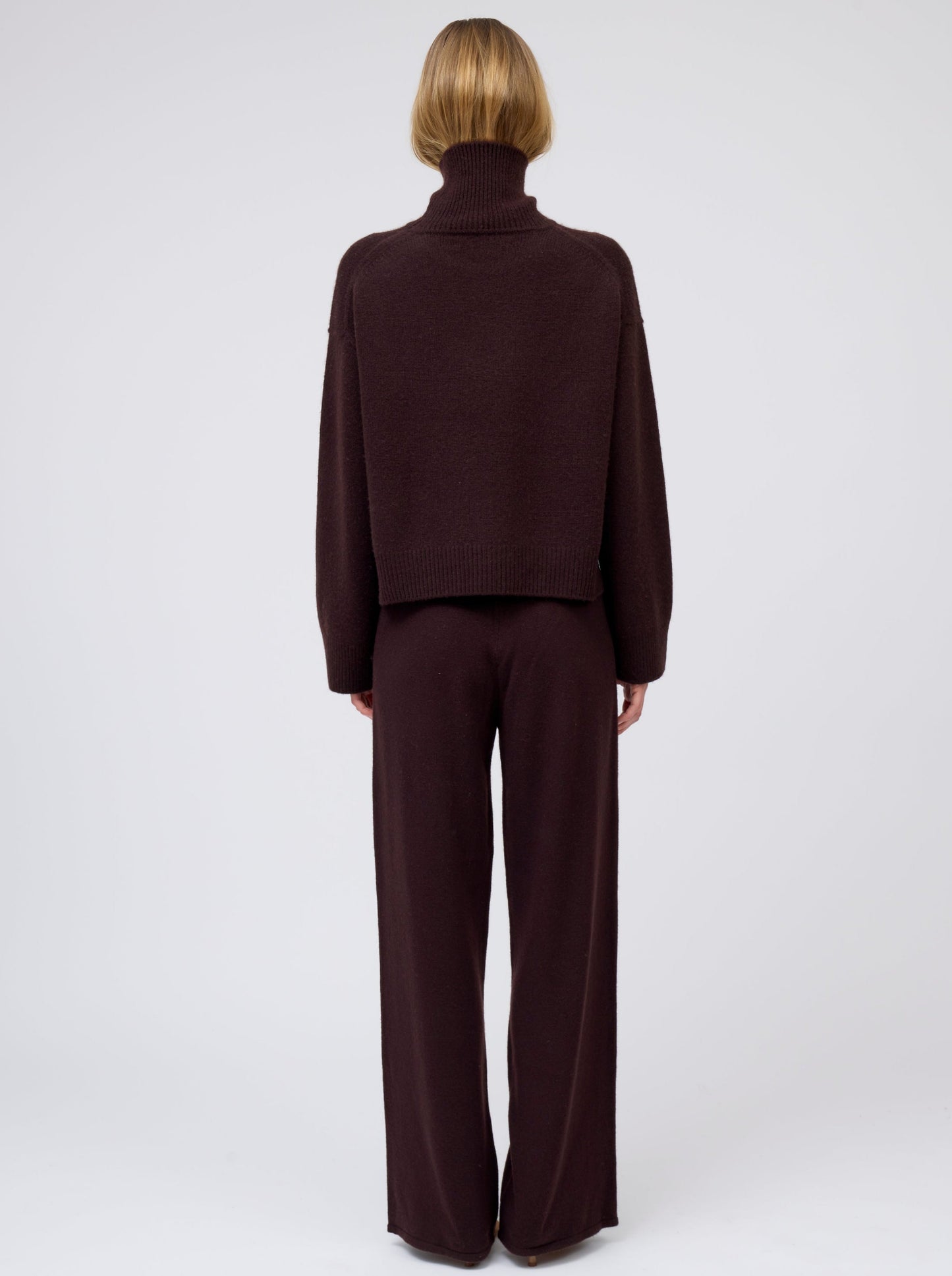 Back view of model wearing mahogany brown cashmere cropped length roll neck sweater with generous long sleeves; worn with  matching brown cashmere wide leg pants.