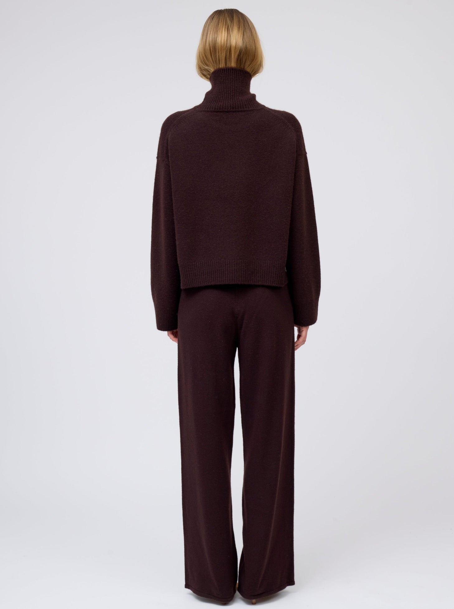 Back view of model wearing mahogany brown cashmere cropped length roll neck sweater with generous long sleeves; worn with  matching brown cashmere wide leg pants.