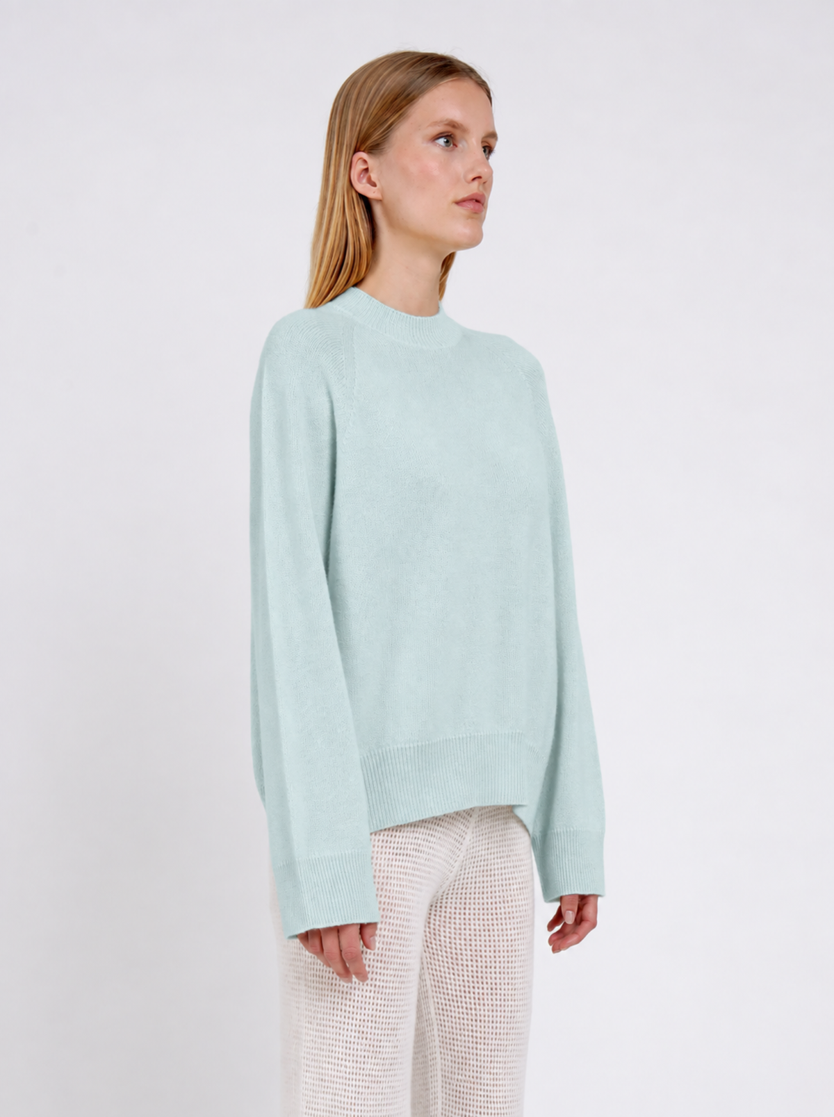 A model stands against a plain white studio background wearing a pale mint green knit sweater and cream-colored textured knit pants.