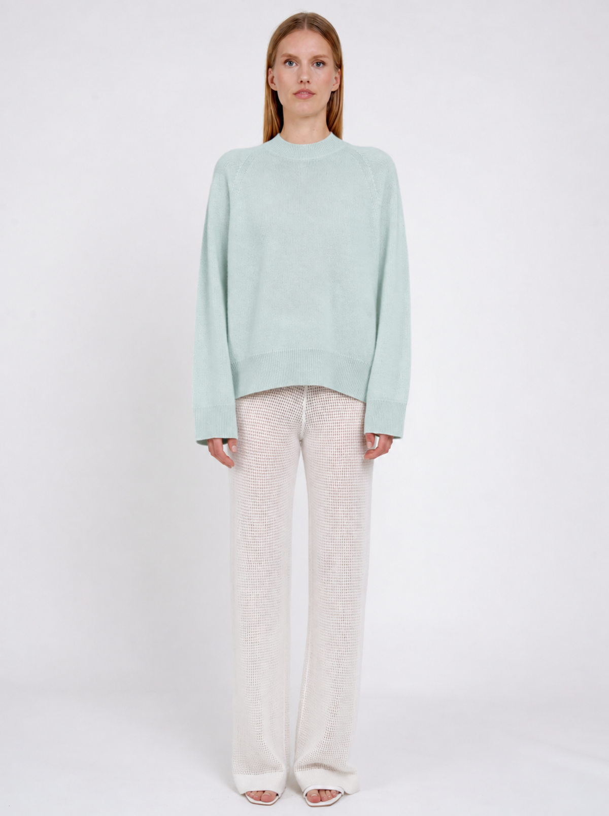 A model stands against a plain white studio background wearing a pale mint green knit sweater and cream-colored textured knit pants.