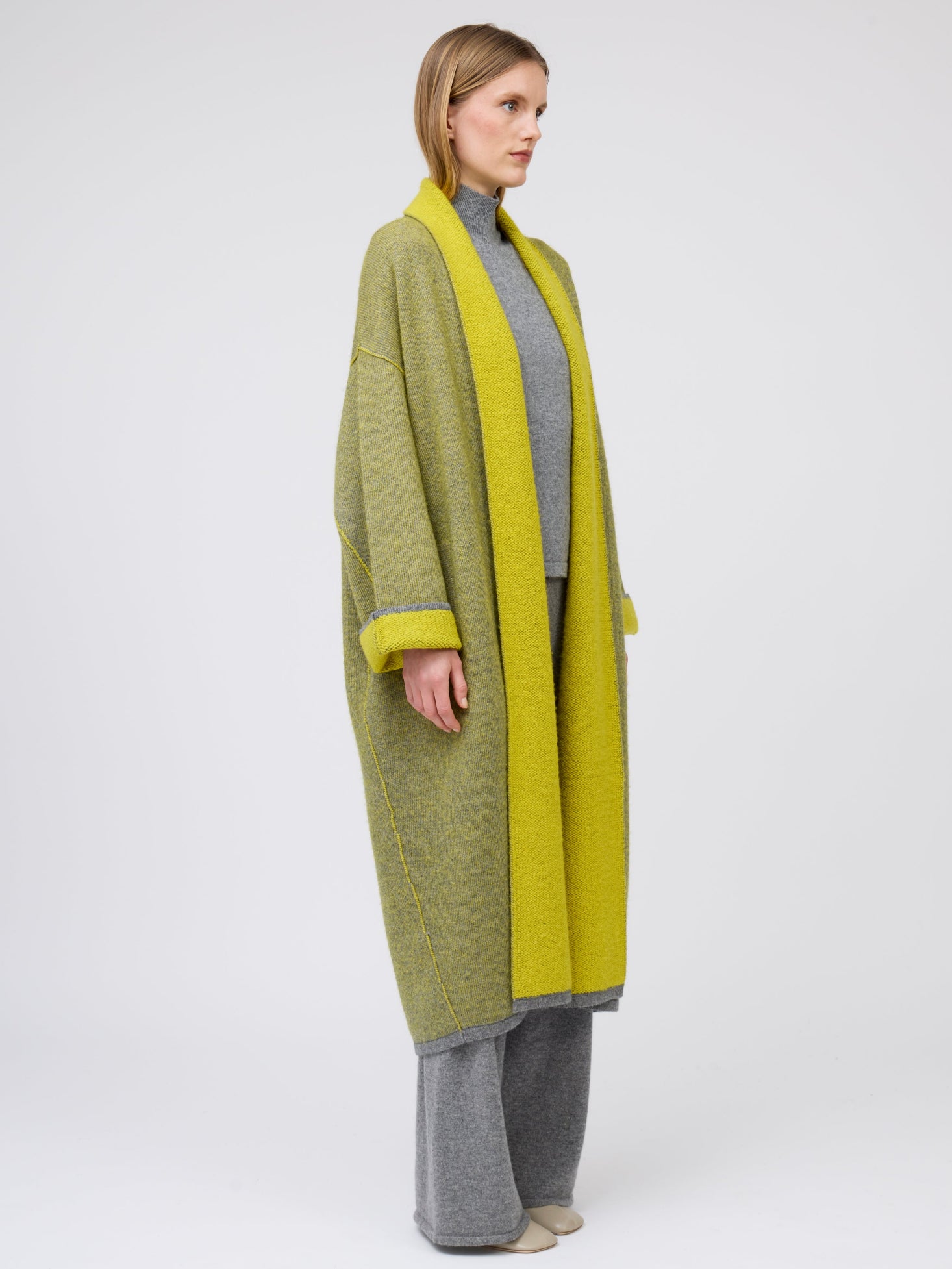 Side view model in long mustard yellow and grey mix cashmere knit coat with yellow collar revere and turn back cuffs; wearing with grey knit turtle neck top and wide leg cashmere pants.