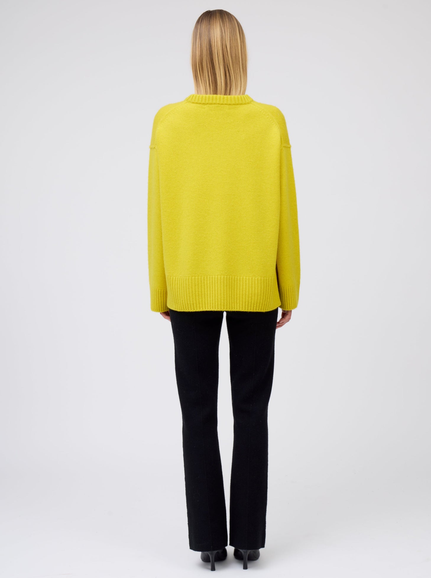 Back view of model wearing mustard yellow oversized round neck sweater with black suit pants and black high heeled boots.