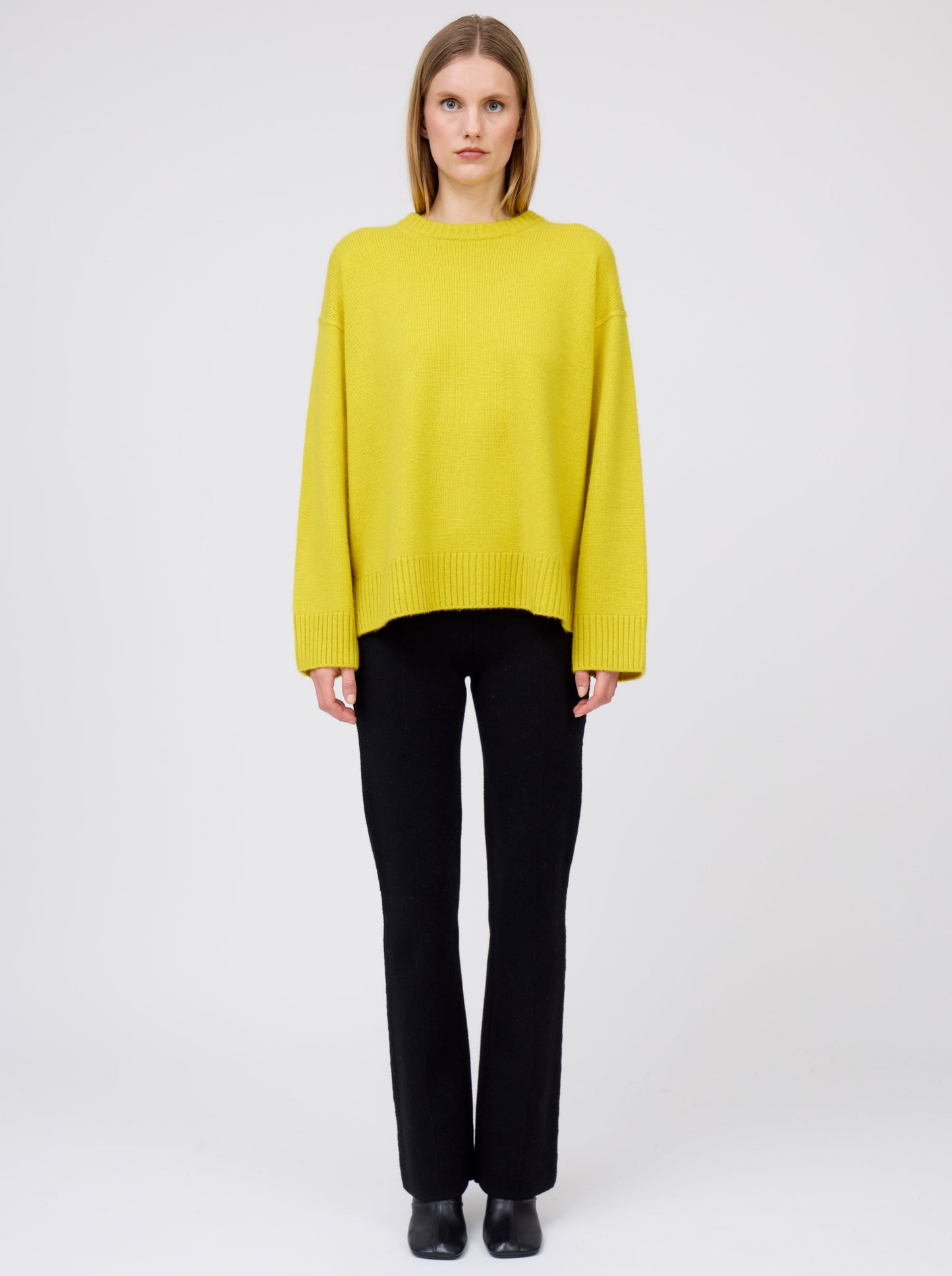 Front view of model in mustard yellow oversized round neck sweater with black suit pants and black boots.