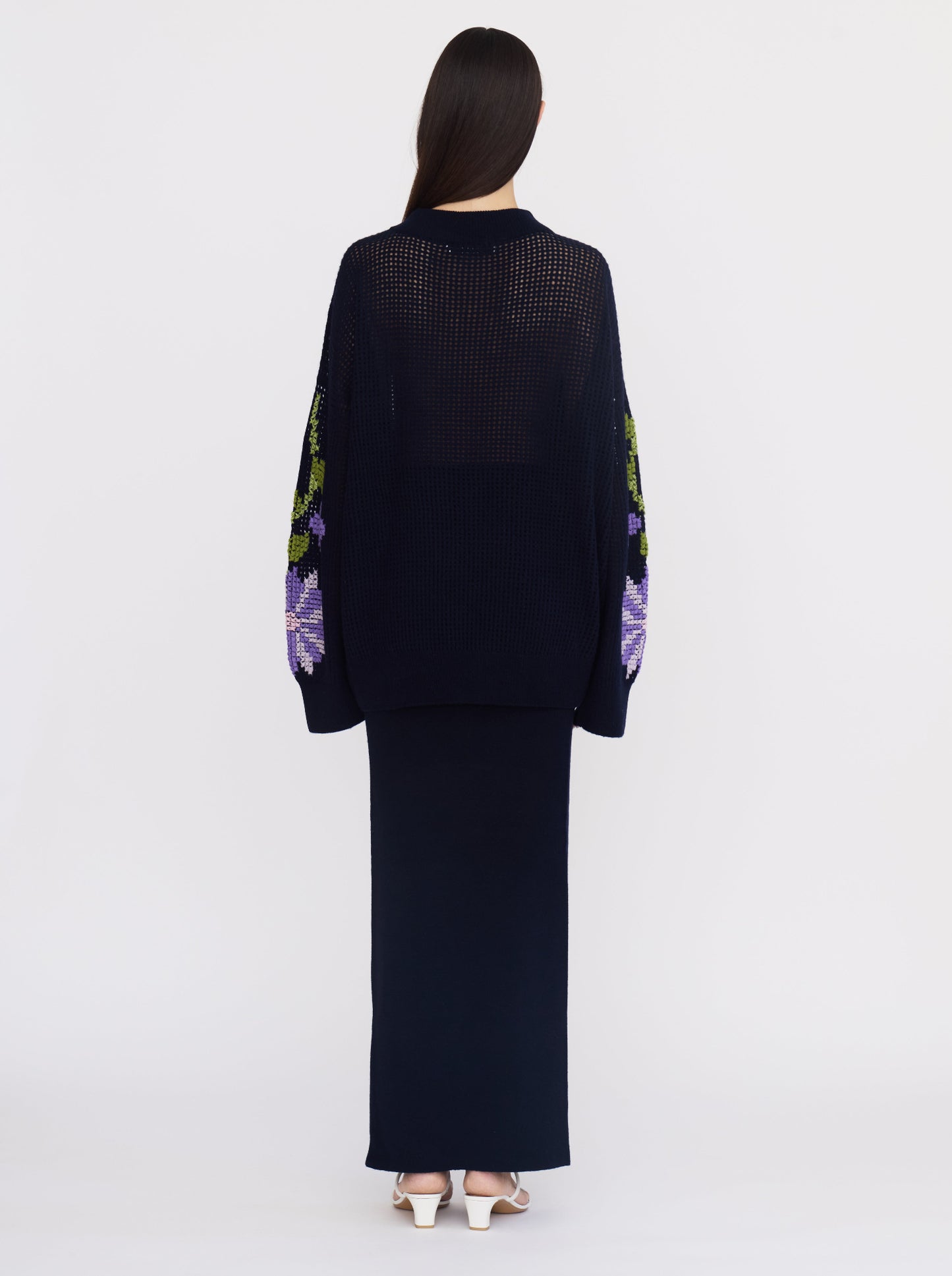 Back view of model wearing dark navy net-knit cashmere oversize jumper with hand-embroidery on the sleeves; worn with a maxi-length dark navy skirt.