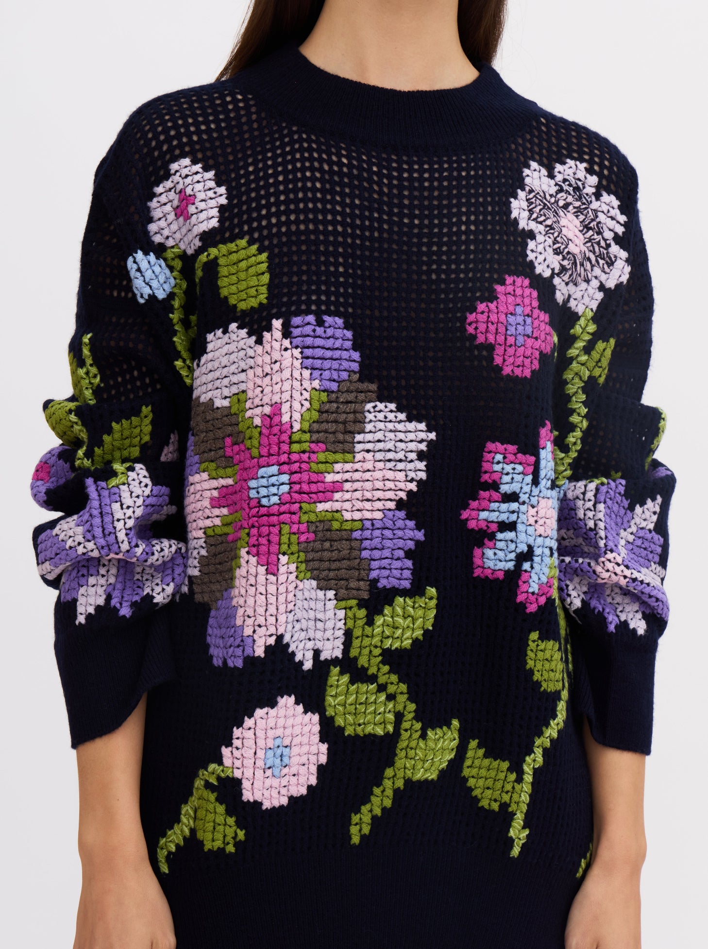 Close-up of model wearing dark navy net-knit cashmere oversize round neck jumper embellished with hand-embroidered cross-stitch floral pattern in multi-coloured cashmere yarns.