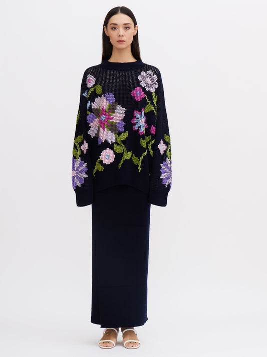 Front view of model wearing dark navy net-knit cashmere oversize jumper embellished with hand-embroidered cross-stitch floral pattern in multi-coloured cashmere yarns; worn with a maxi dark navy skirt.