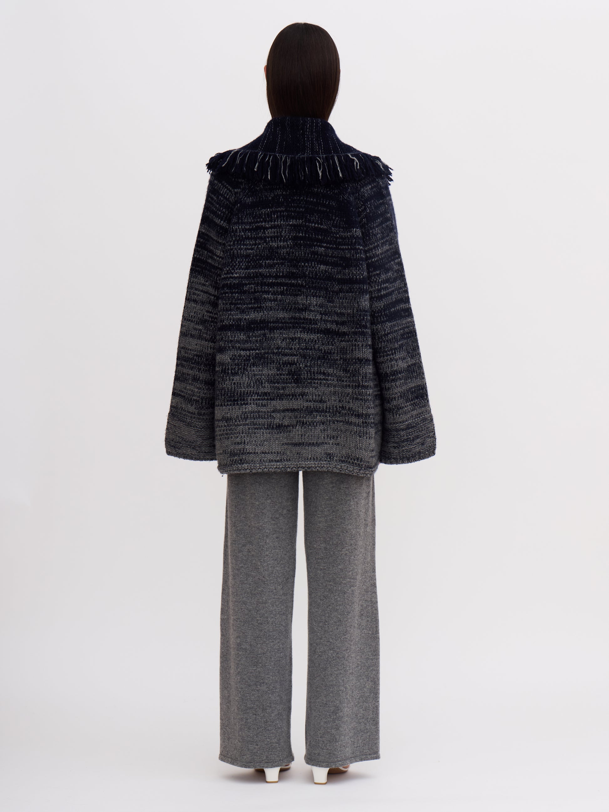 Back view of model wearing chunky cashmere knit jacket with fringed shawl collar and grey cashmere pants.