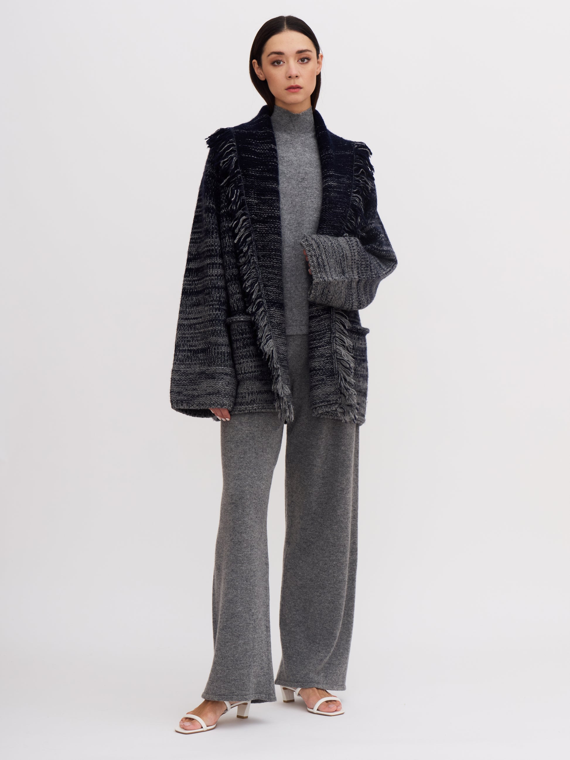 Model wearing chunky knit cashmere hip length jacket with fringed shawl collar and grey wide leg cashmere pants, showing wide jacket sleeves and deep cuffs. 