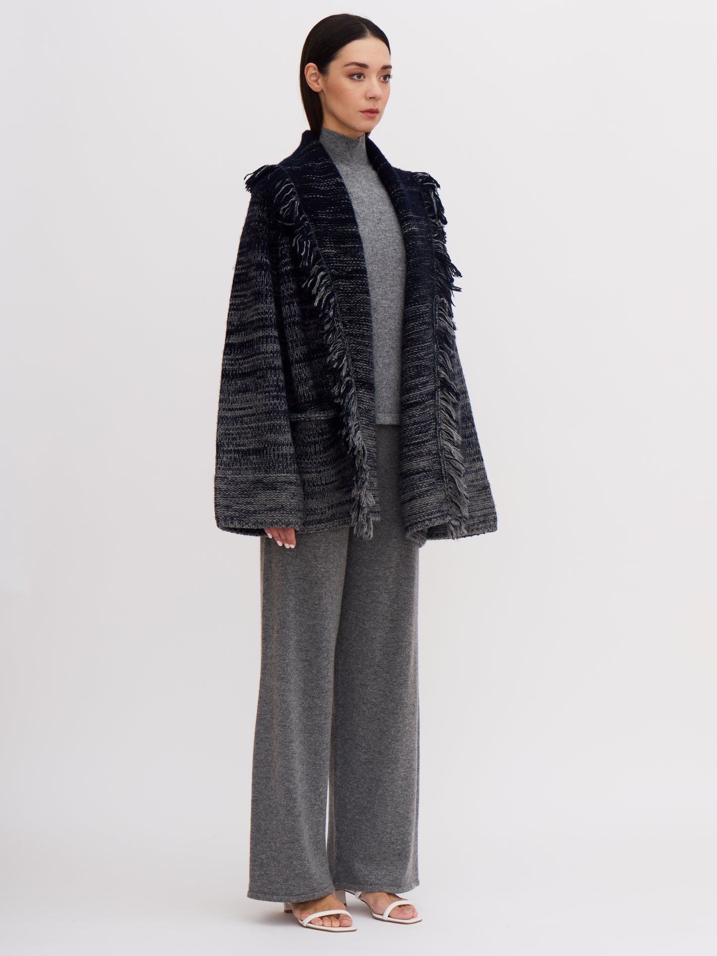 Model wearing chunky cashmere knit jacket in navy and grey mix, with fringed shawl collar and grey wide leg cashmere pants.