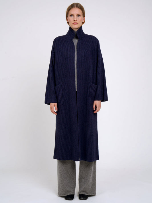 Model wearing a long navy cardigan coat in ribbed knit cashmere with high collar, patch pockets and generous long sleeves; worn with grey cashmere pants.