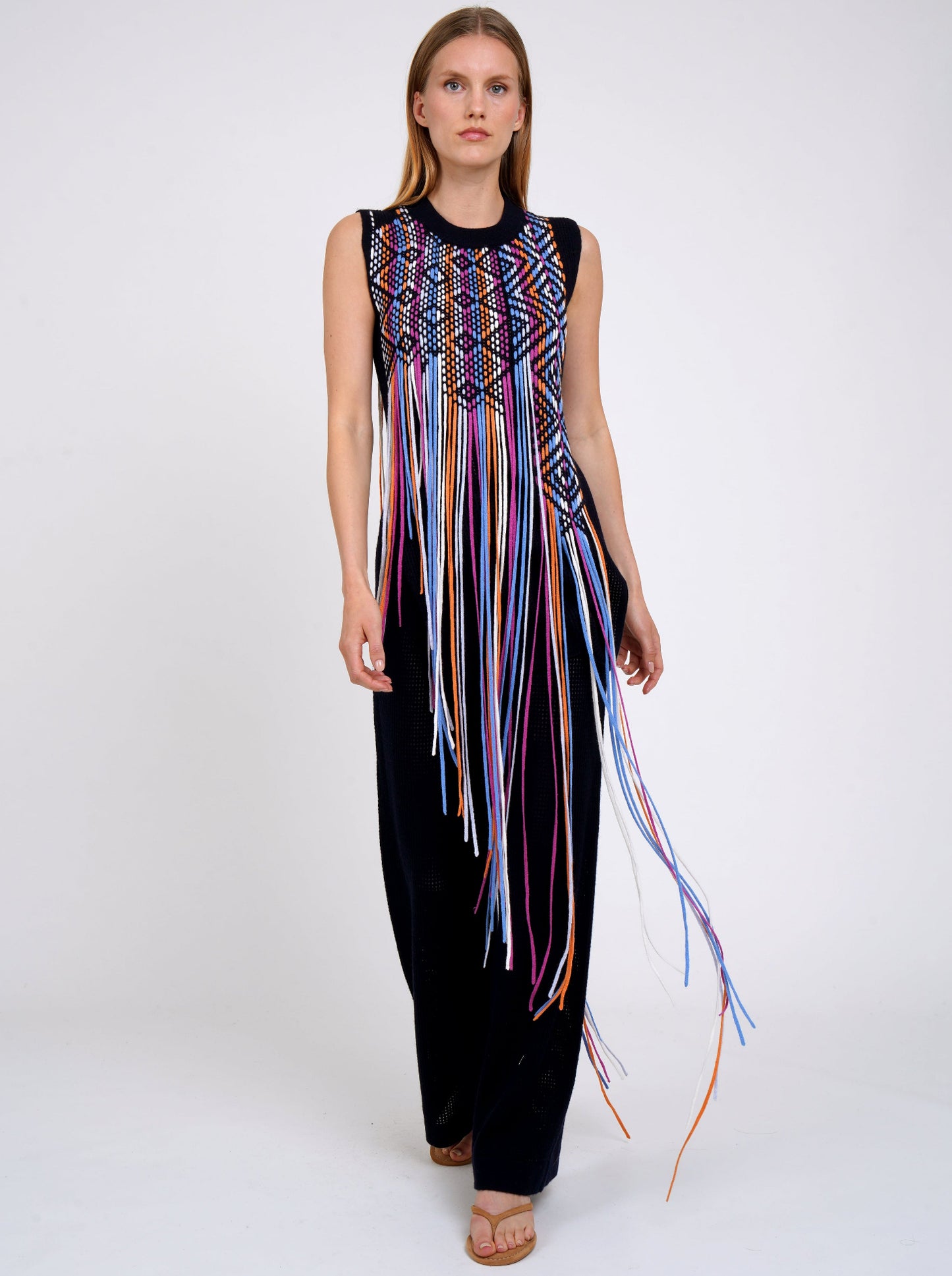model walks towards camera wearing Navy light weight cashmere sleeveless tunic decorated with woven coloured long fringes worn with navy knit wide leg pants.
