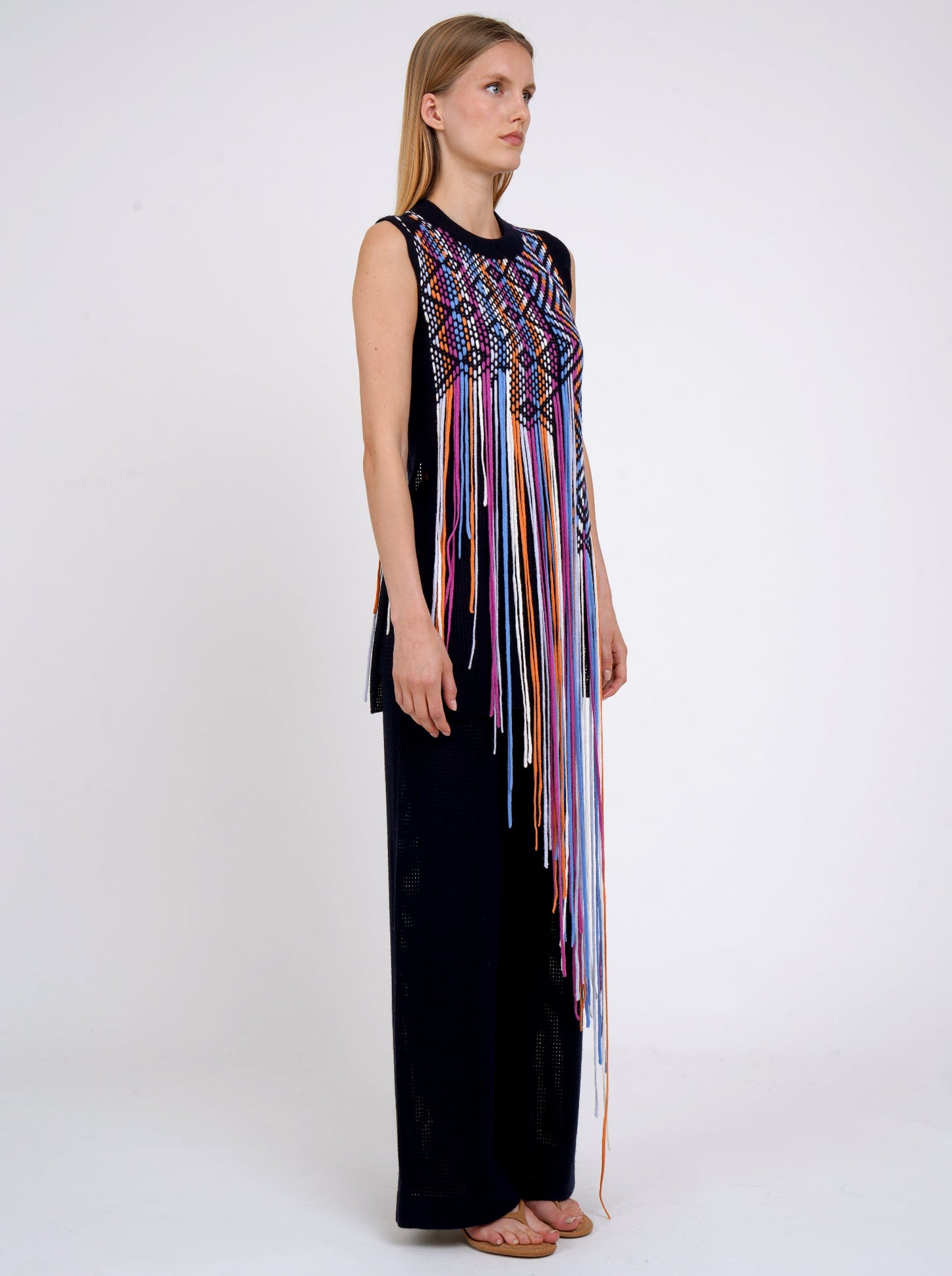 Side view model in navy light weight cashmere sleeveless tunic decorated with woven coloured long fringes worn with navy knit wide leg pants.