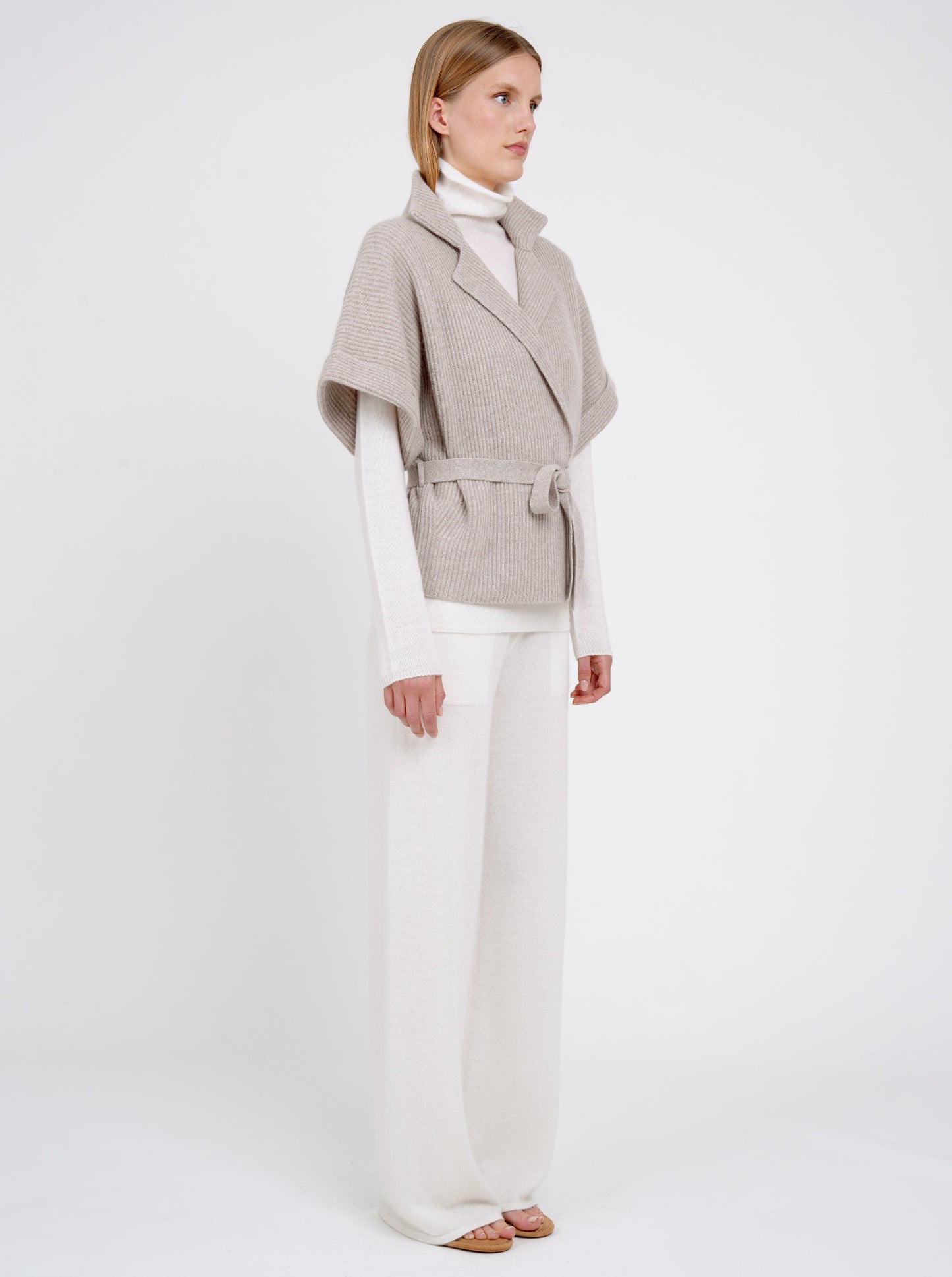 Side view model wears beige cashmere short-sleeve ribbed knit short jacket over white knit turtle neck top and white wide leg pants.