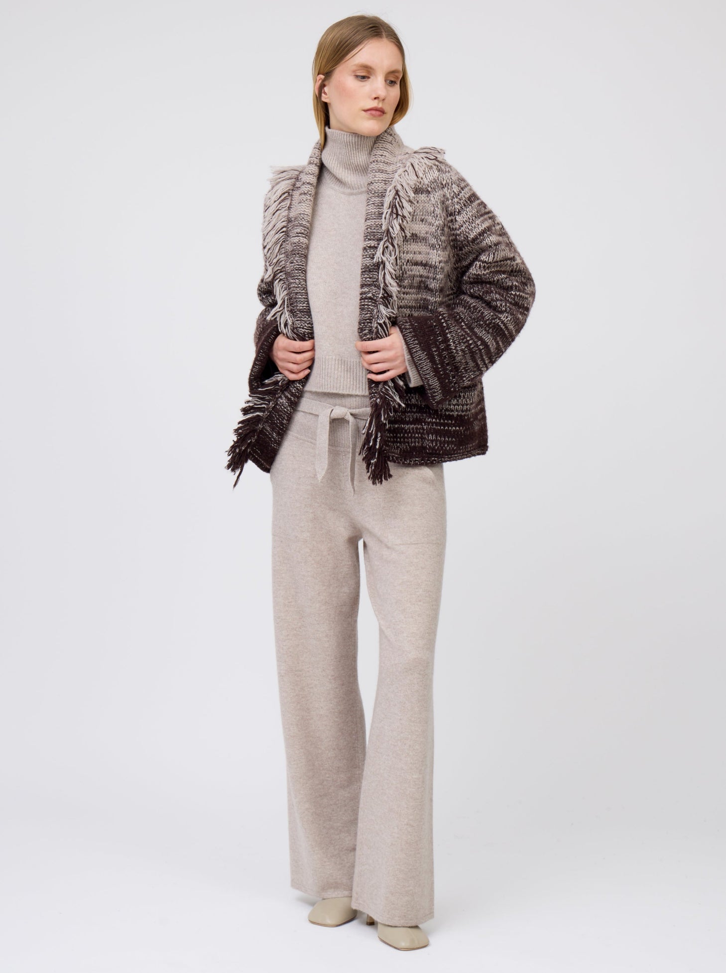 Model wears beige cashmere, belted, wide leg pants with pockets; worn with knitted brown and beige mix short jacket with fringe collar over cropped, roll neck, beige sweater.
