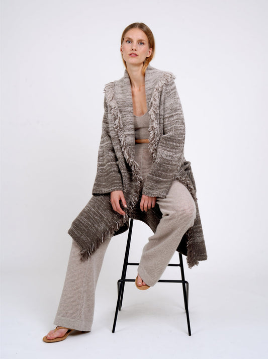 Sitting in relaxed pose, model wears mixed colour knitted coat in beige and brown with fringed shawl collar, long wide sleeves; with beige cashmere knit pants.